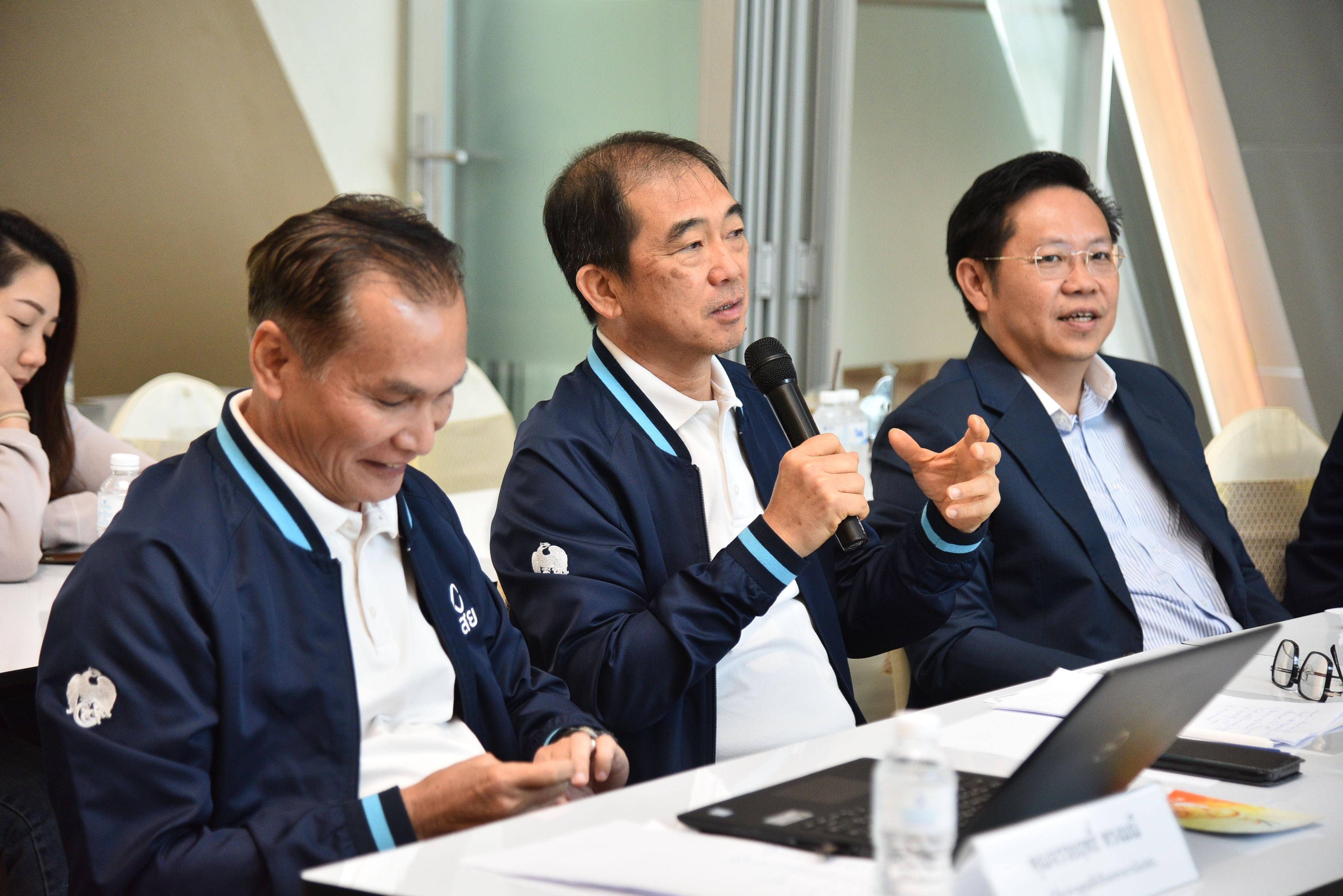 Three men in business casual attire attend a meeting, one speaking into a microphone.