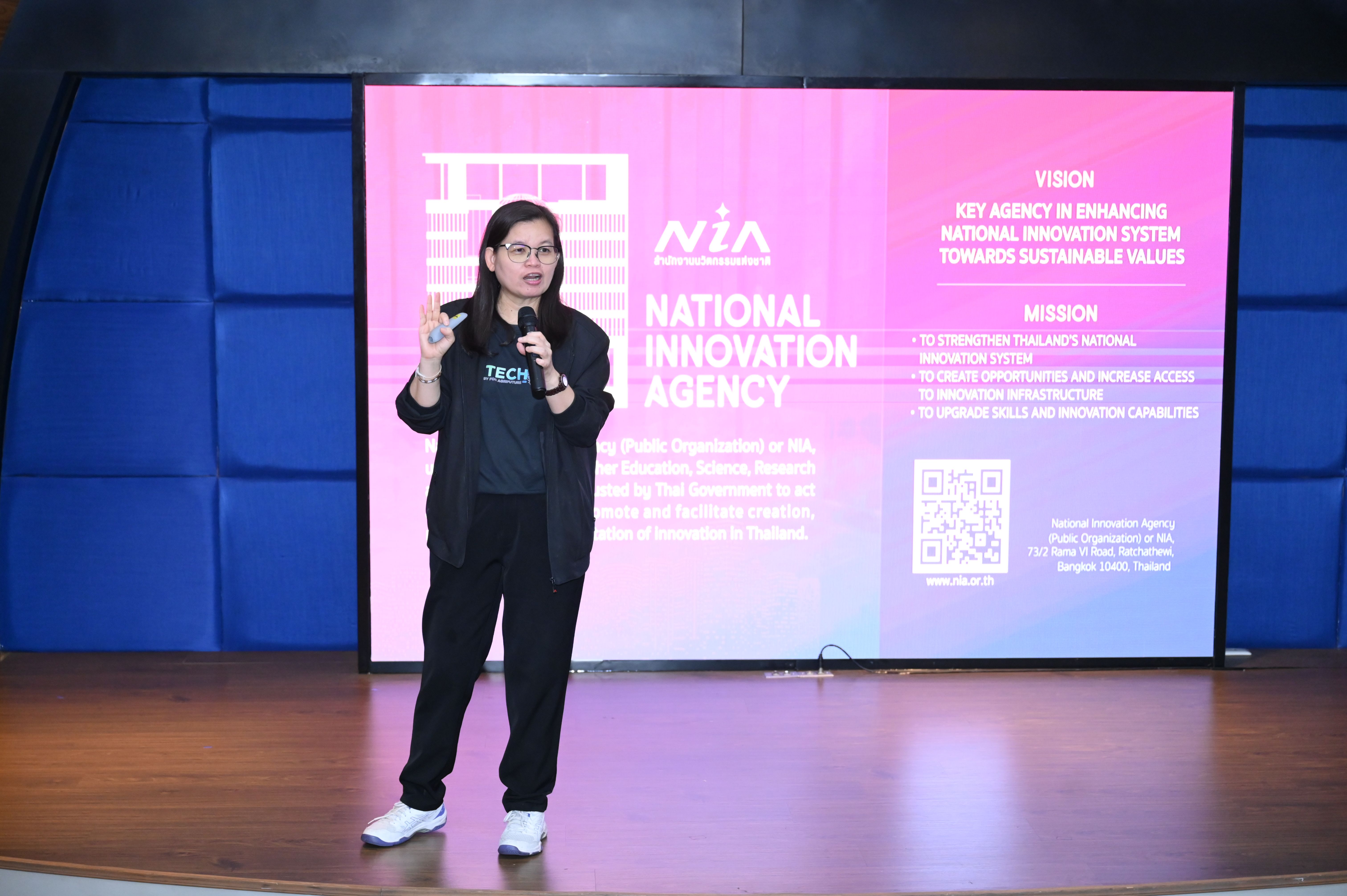 Woman speaks into microphone on stage in front of a screen displaying "National Innovation Agency."