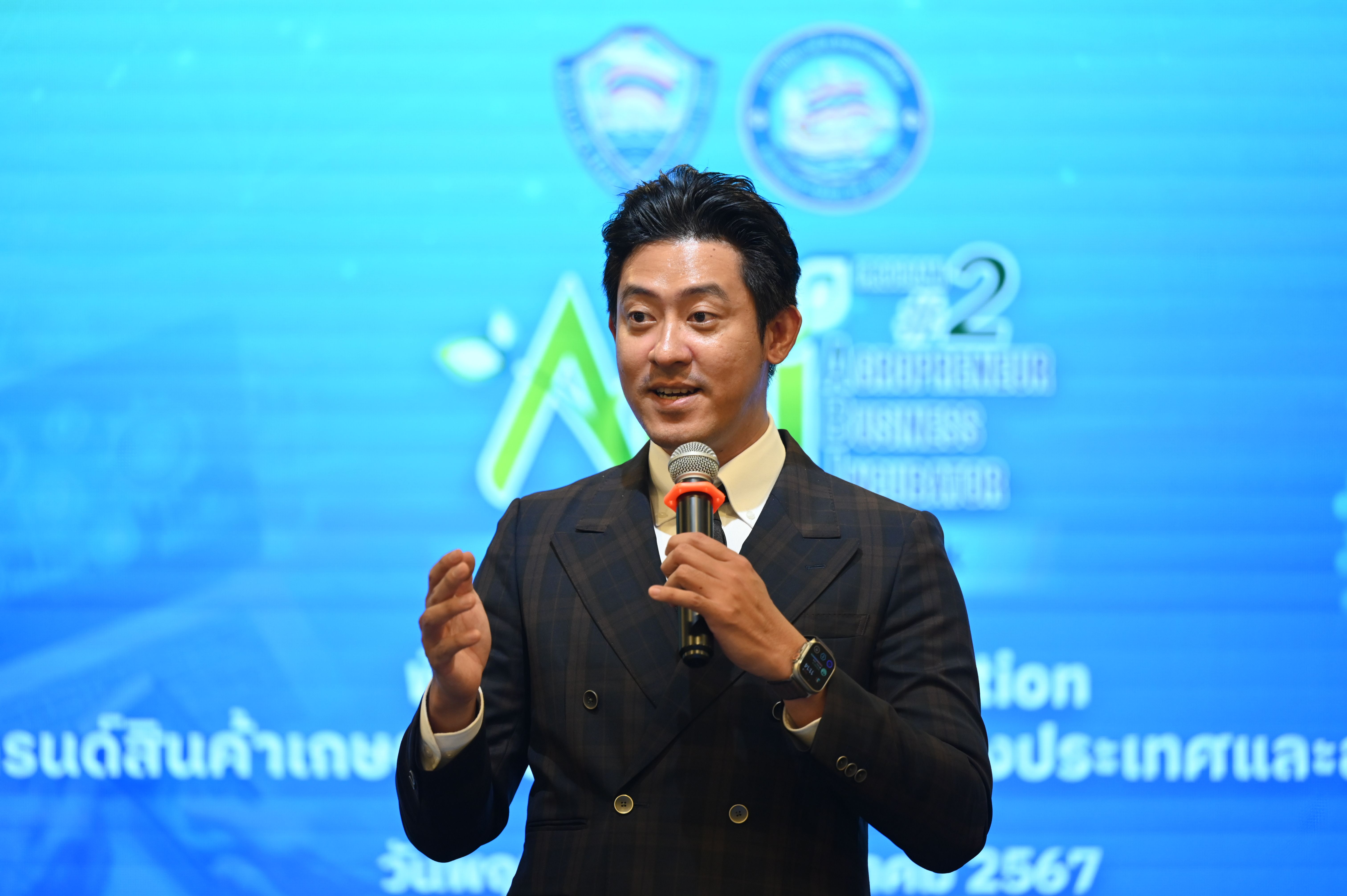 A professional man in a dark suit speaks into a microphone against a blue event background.