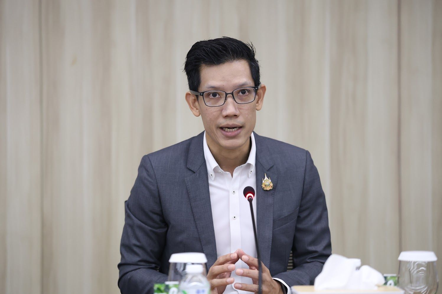 A man in a dark suit and glasses speaks into a microphone at a formal meeting.