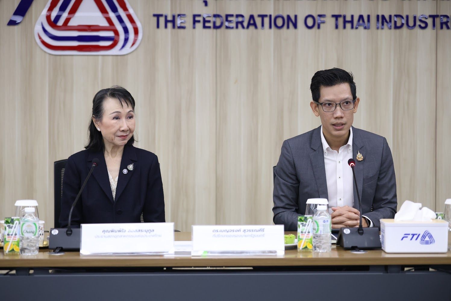 A woman and a man sit at a table with The Federation of Thai Industry sign.