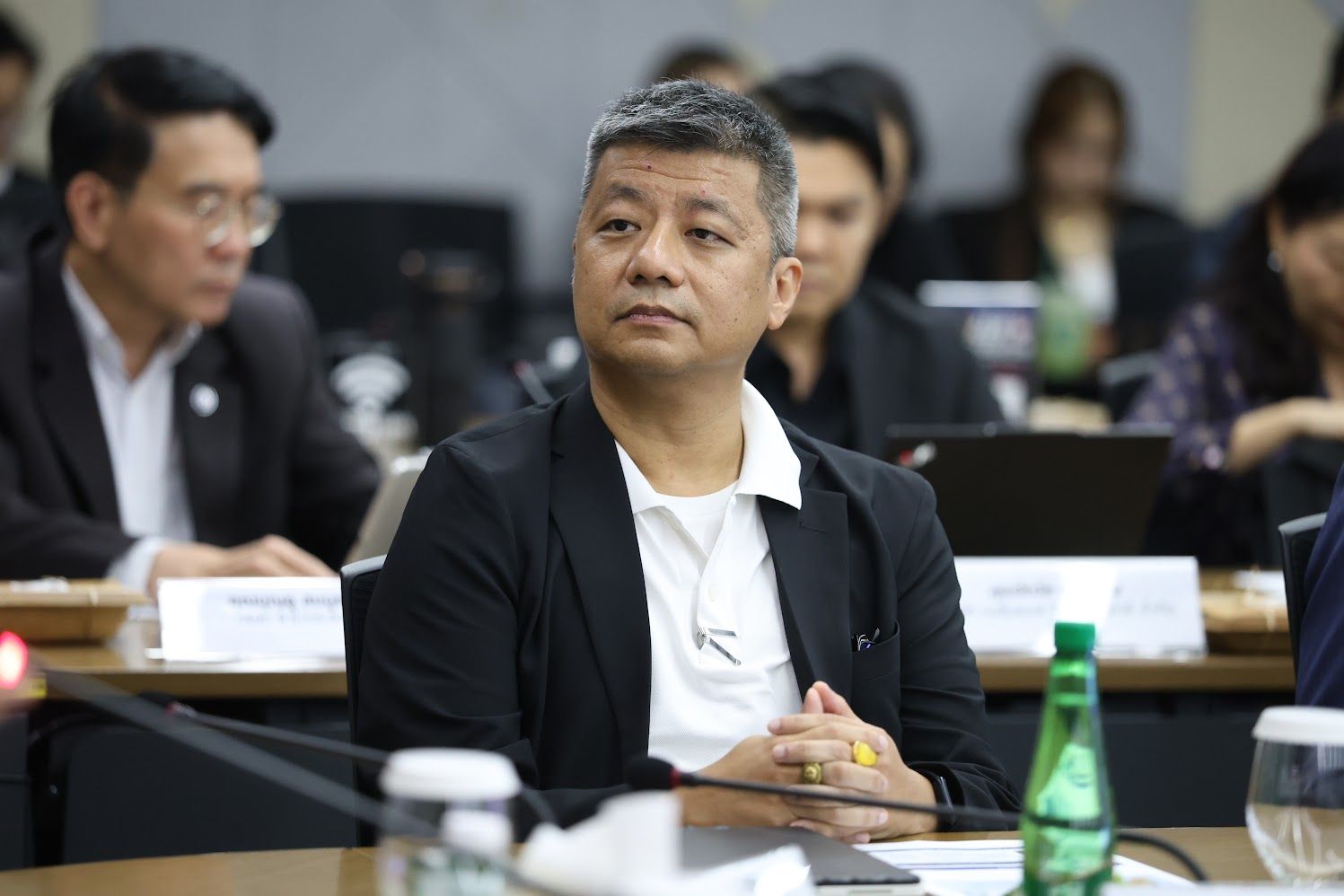 Man in black jacket and white polo sits at conference table, looking attentive.