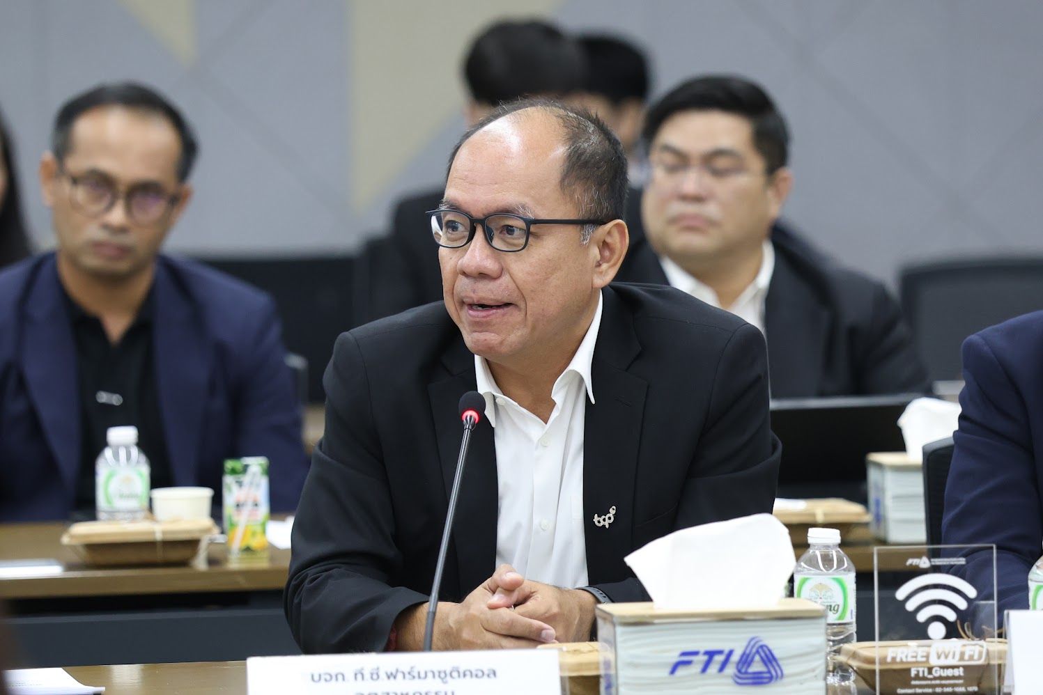 An Asian man in glasses and a suit speaks at a formal meeting table.