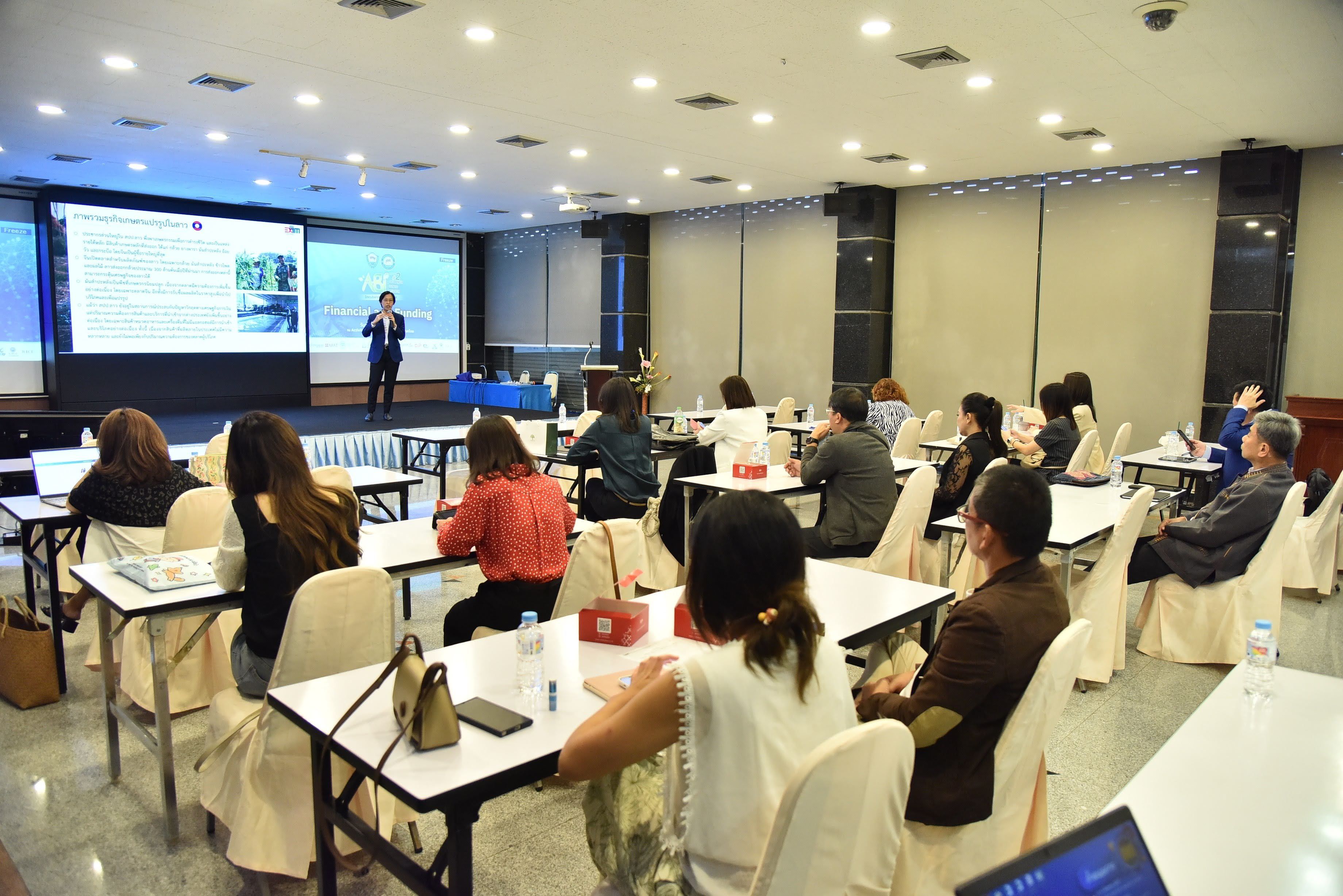 Speaker presents to an audience seated at tables in a brightly lit conference room.