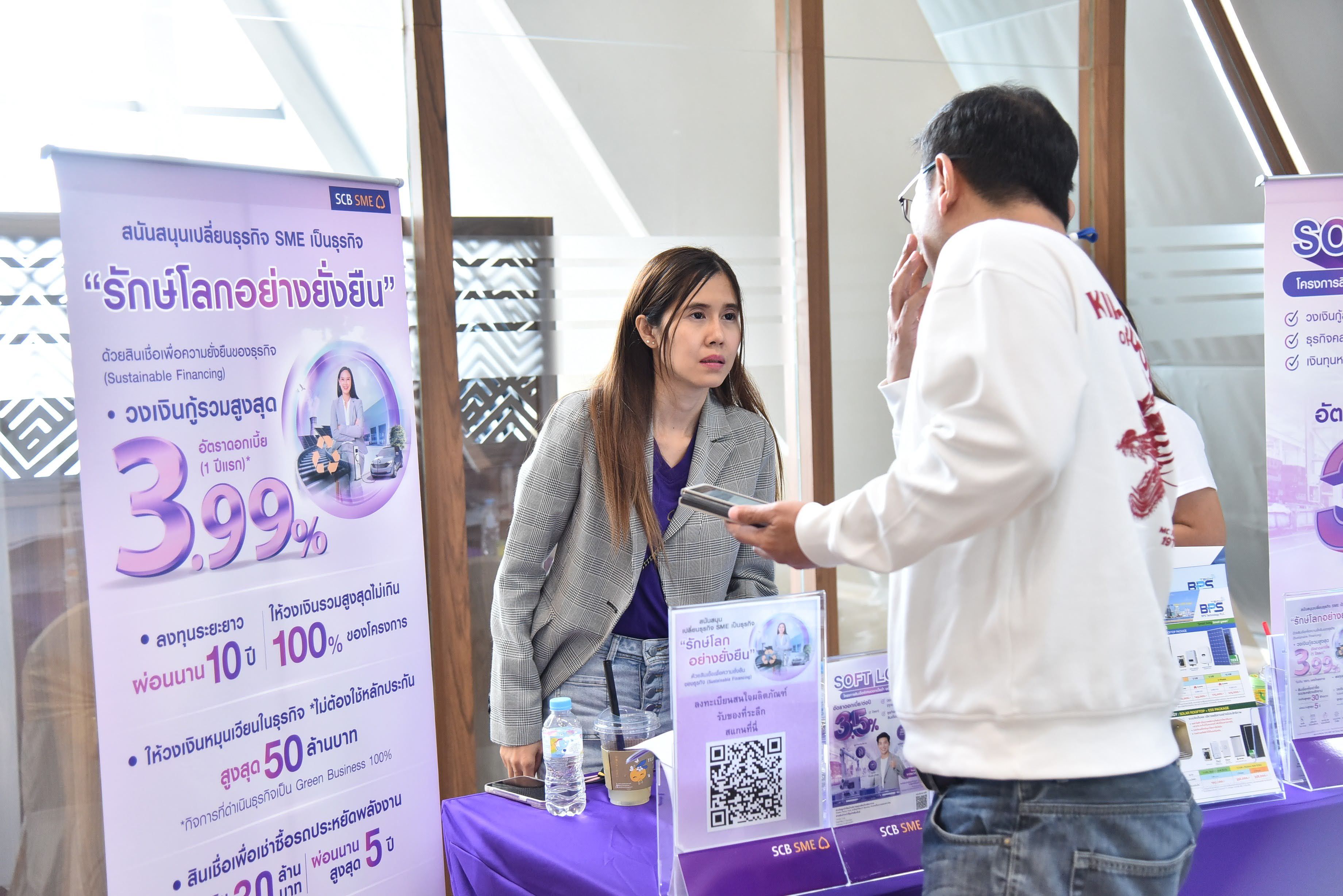 A woman in a grey blazer talks to a man at a purple promotional booth with Thai text.