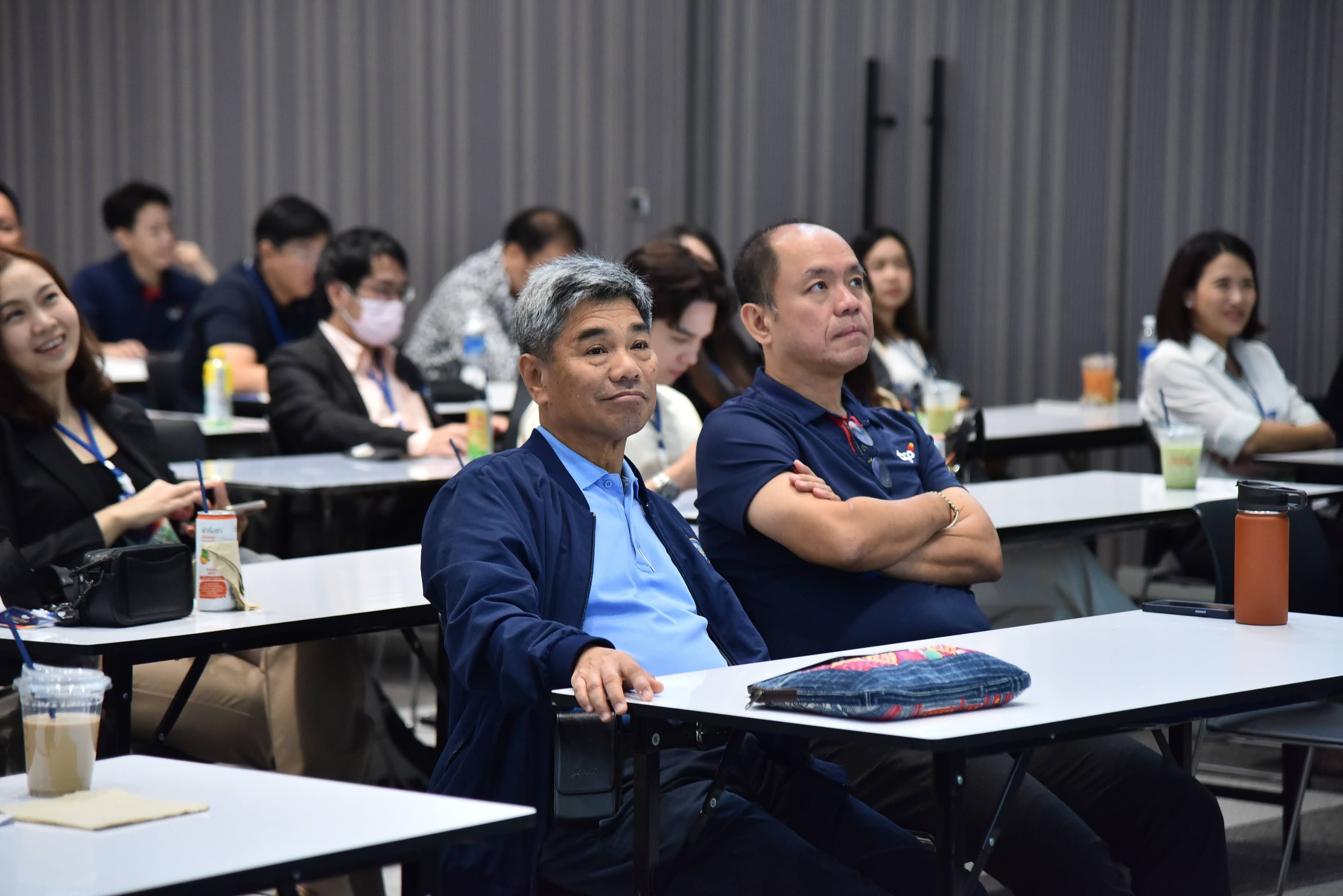 A group of attentive adults sits at desks in a conference room, listening to a speaker.