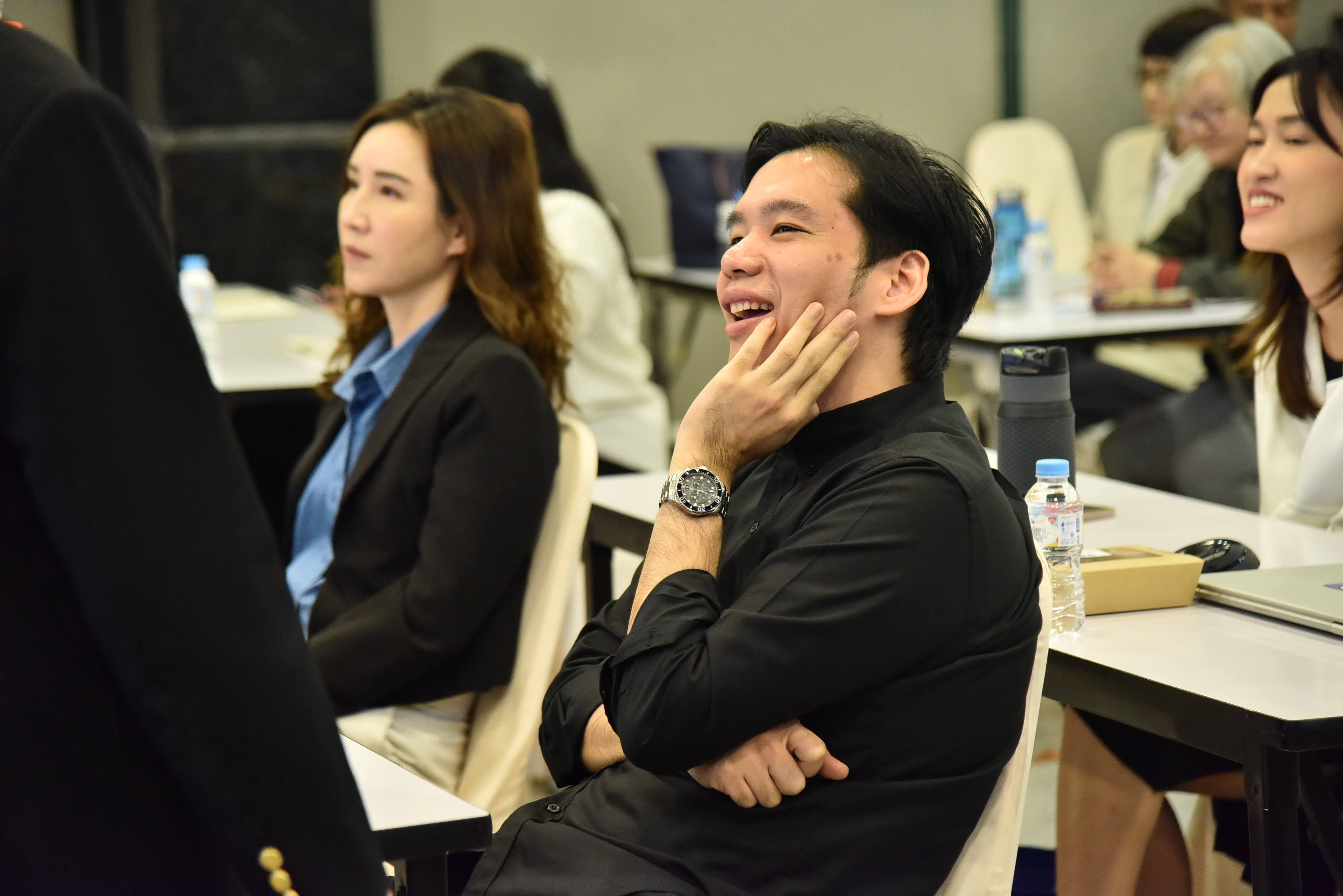 A smiling man in a black shirt rests his chin on his hand at a conference.