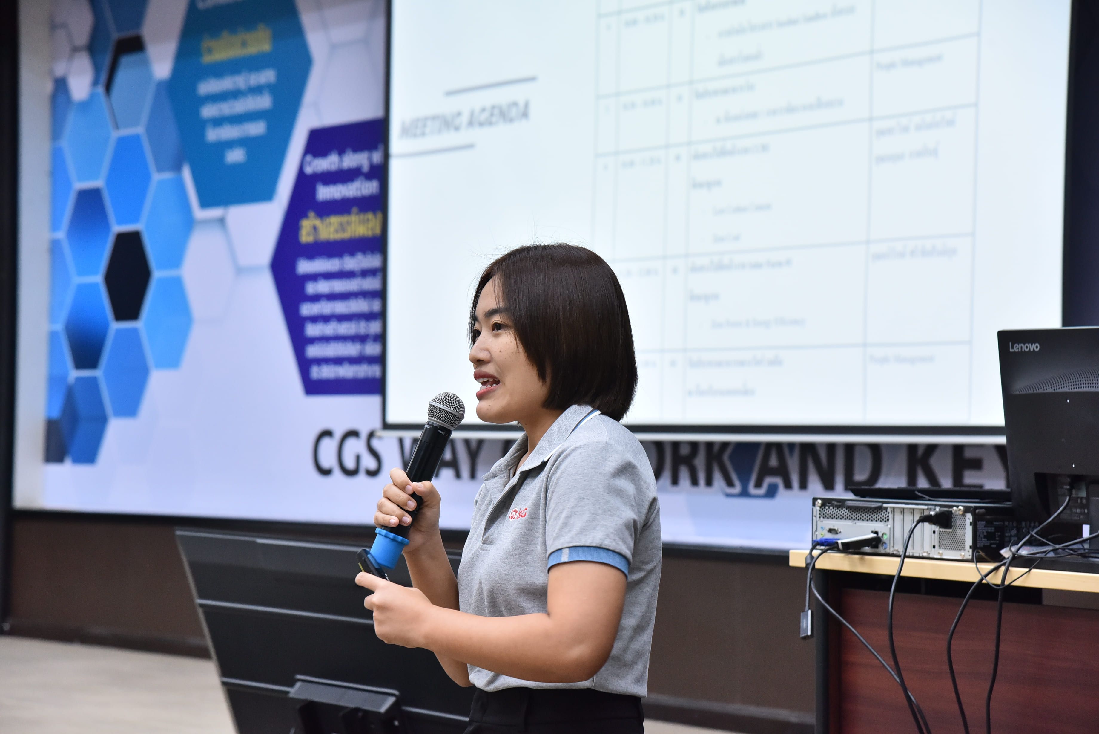 Woman speaking into microphone at a presentation with a meeting agenda on screen.