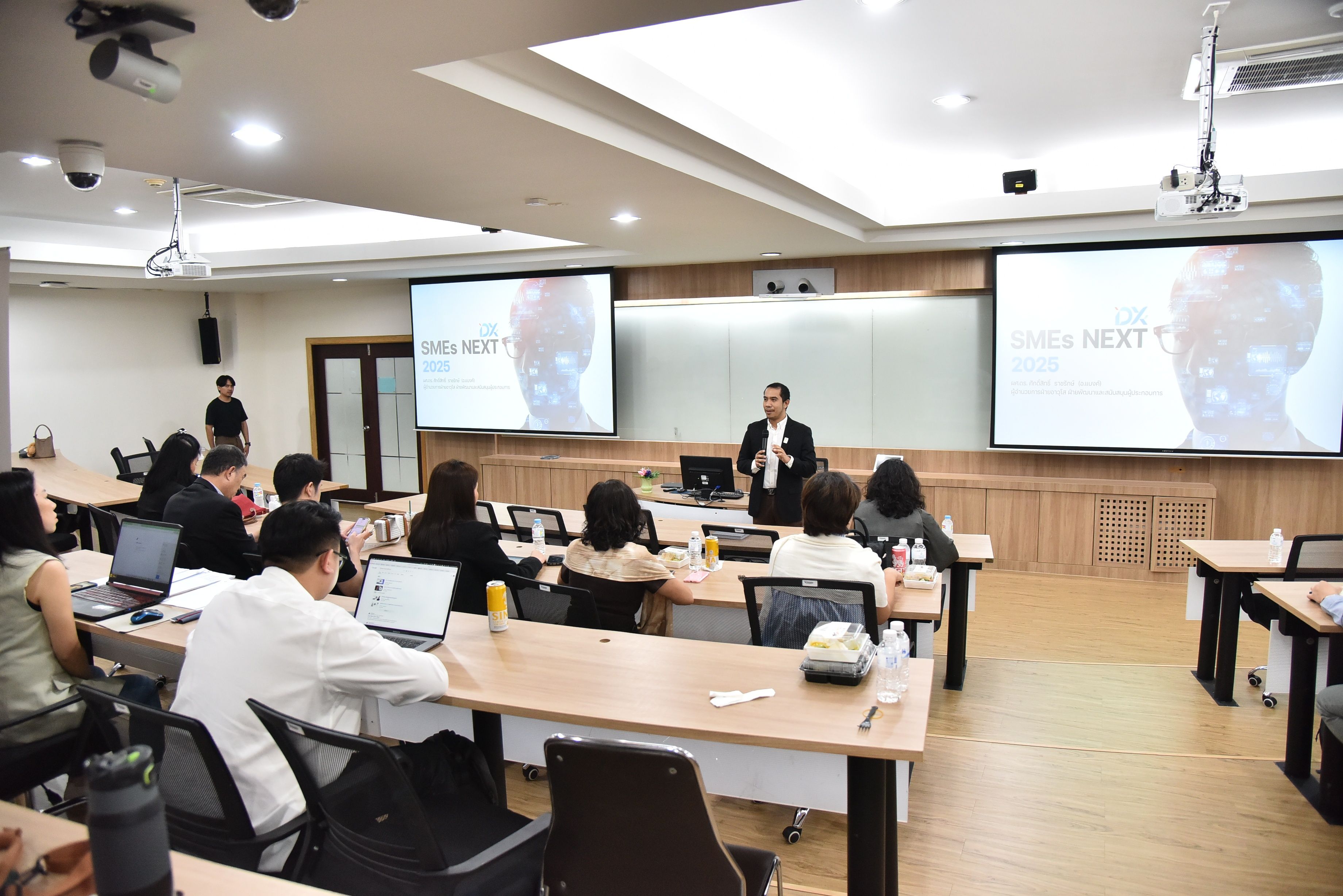 Presenter addresses an audience in a bright classroom with 'WHAT'S NEXT?' on dual screens.