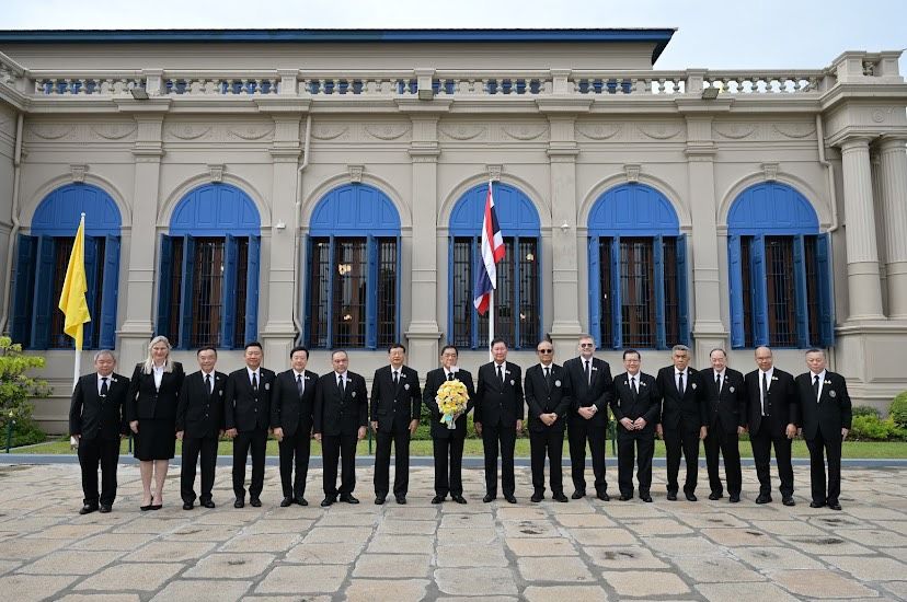 A group of officials in black suits stands formally before a grand building with a Thai flag.