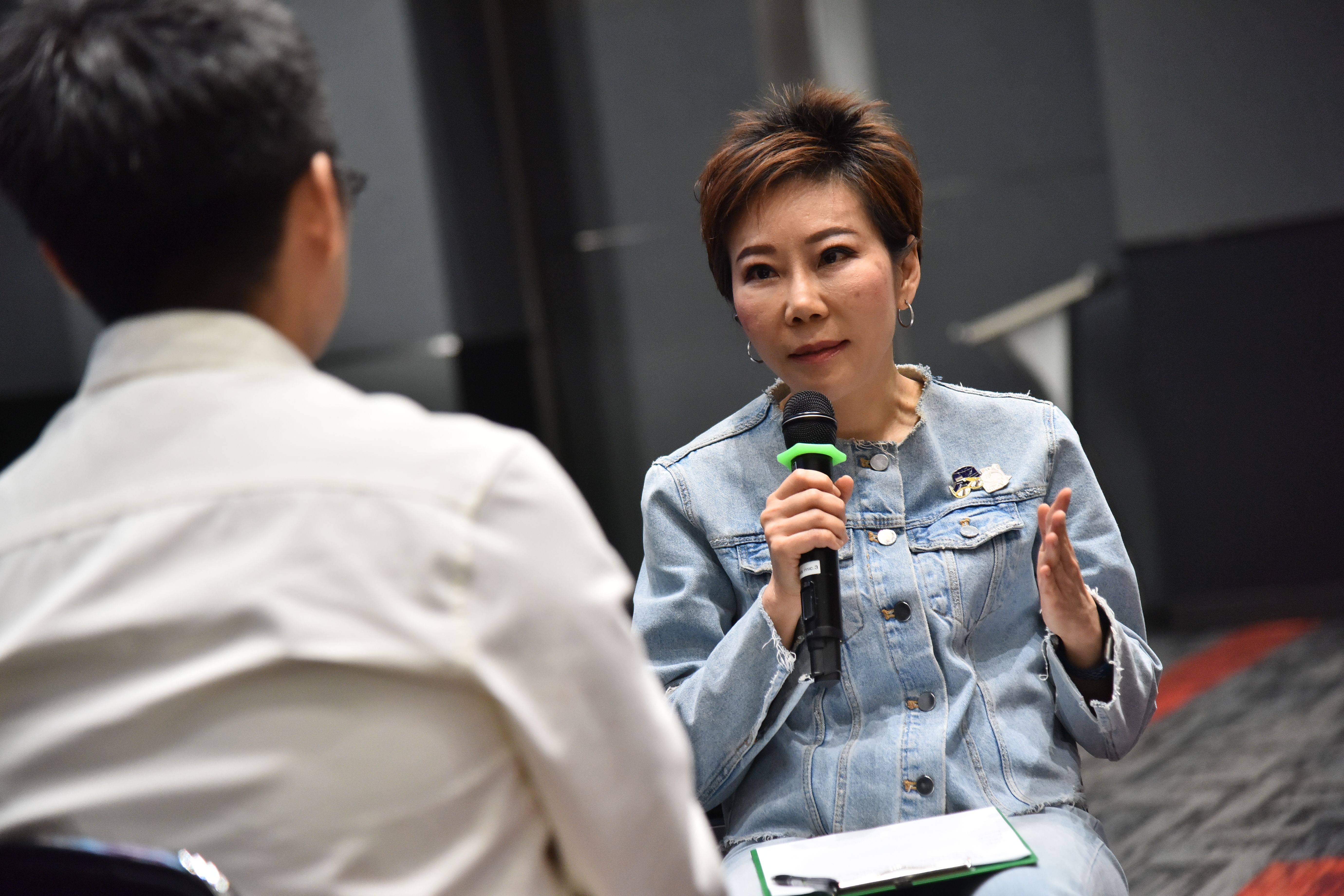Woman in denim jacket speaking into a microphone during an interview.