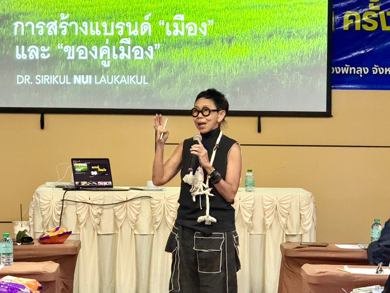 Speaker in glasses and dark sleeveless top gesturing at a podium.