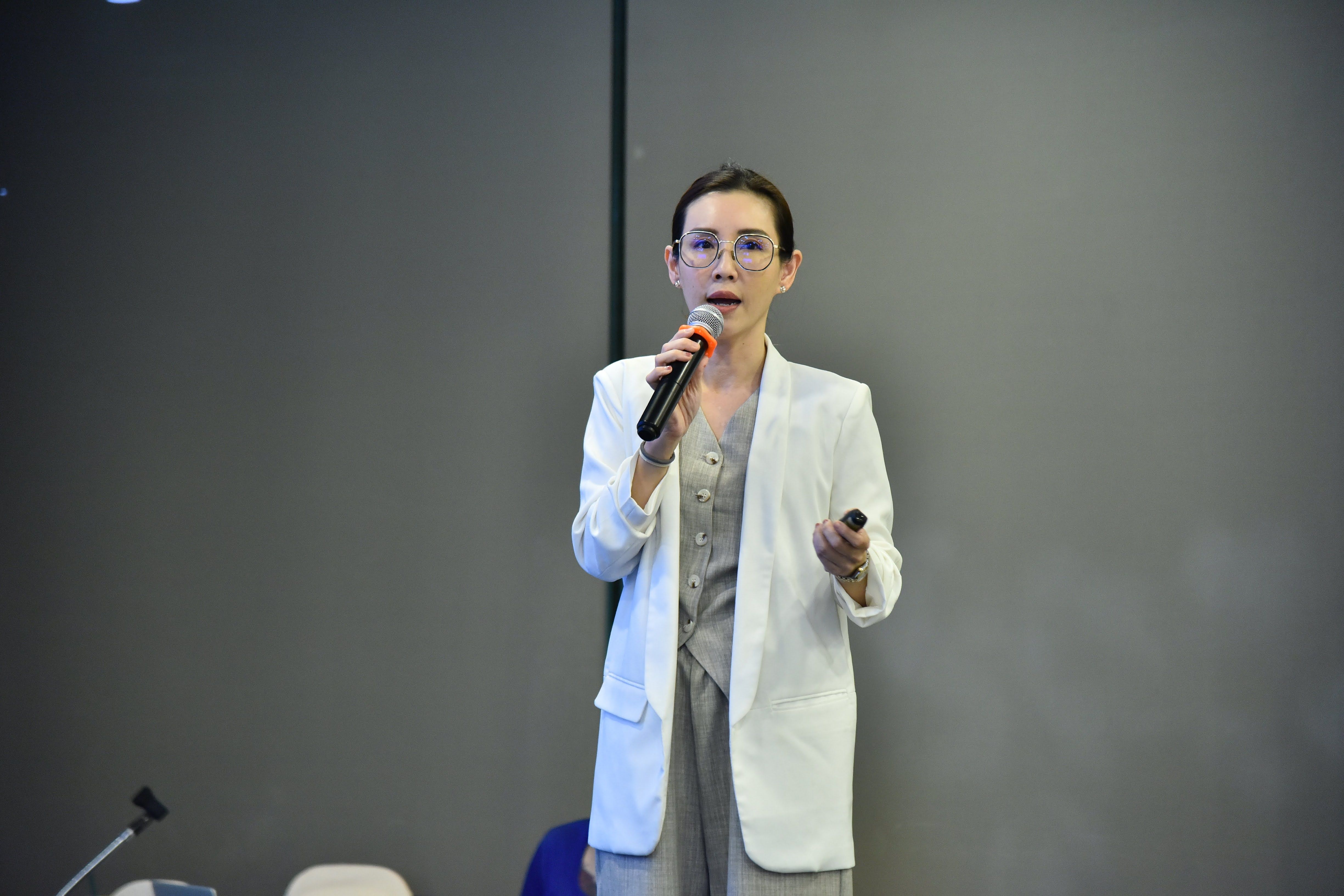 Woman in white blazer speaks into microphone, gesturing during a presentation.