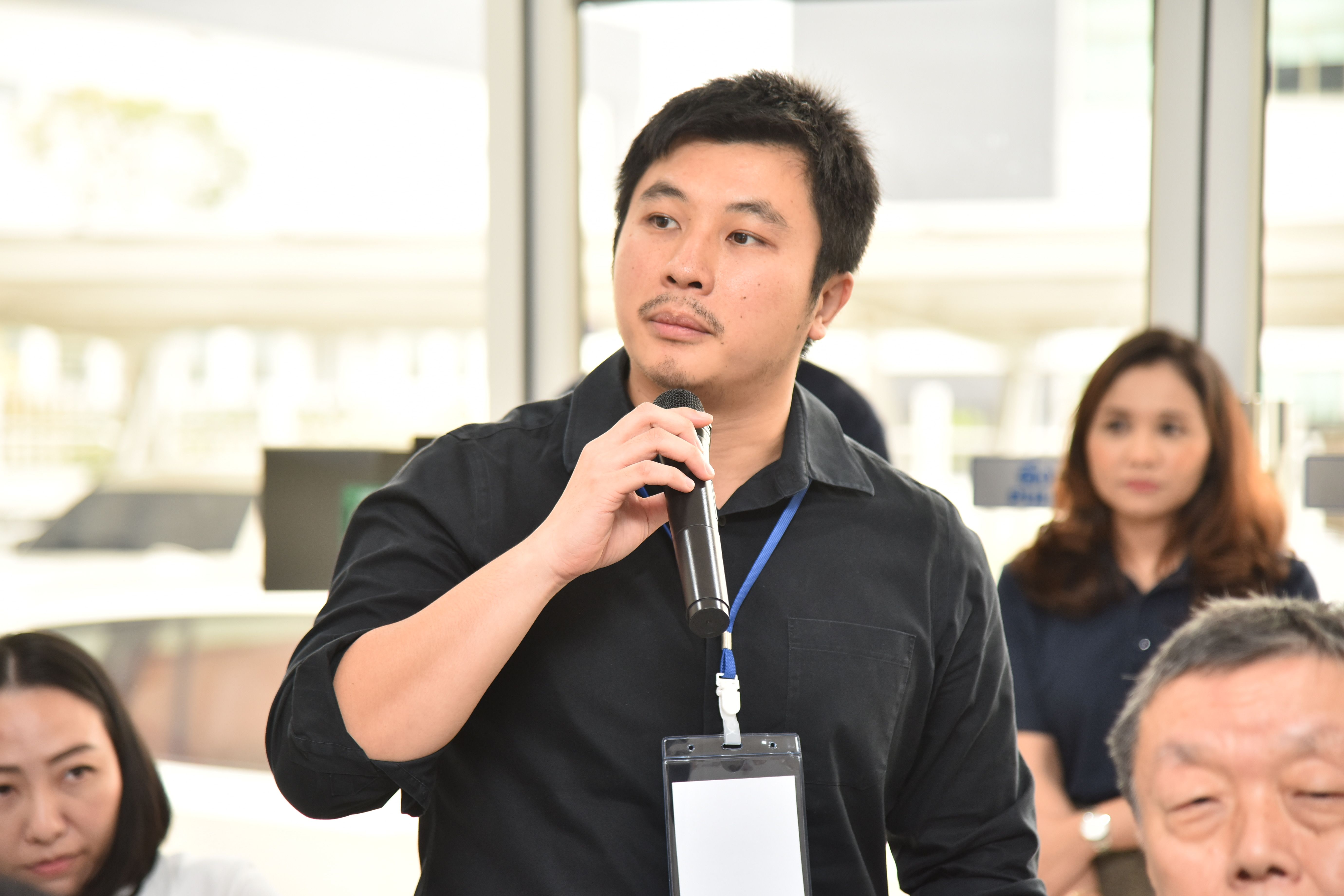 A man in a black shirt speaks into a microphone at an indoor event.