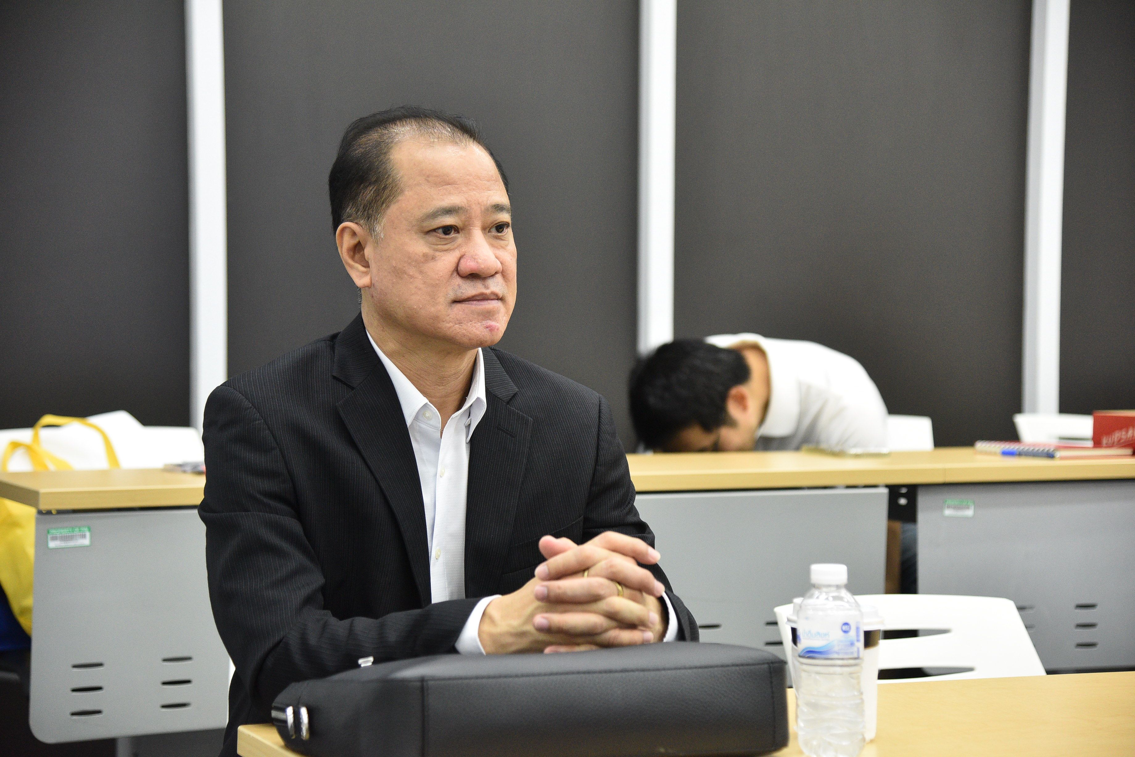 A serious man in a black suit sits at a table, hands clasped, looking right.