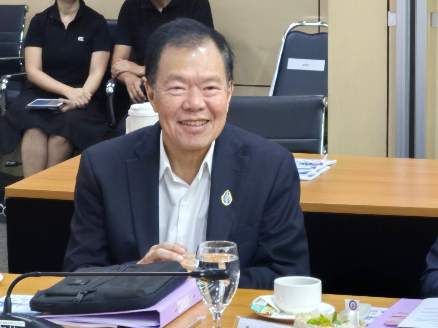 Older Asian man in a dark suit smiling at a conference table with drinks.