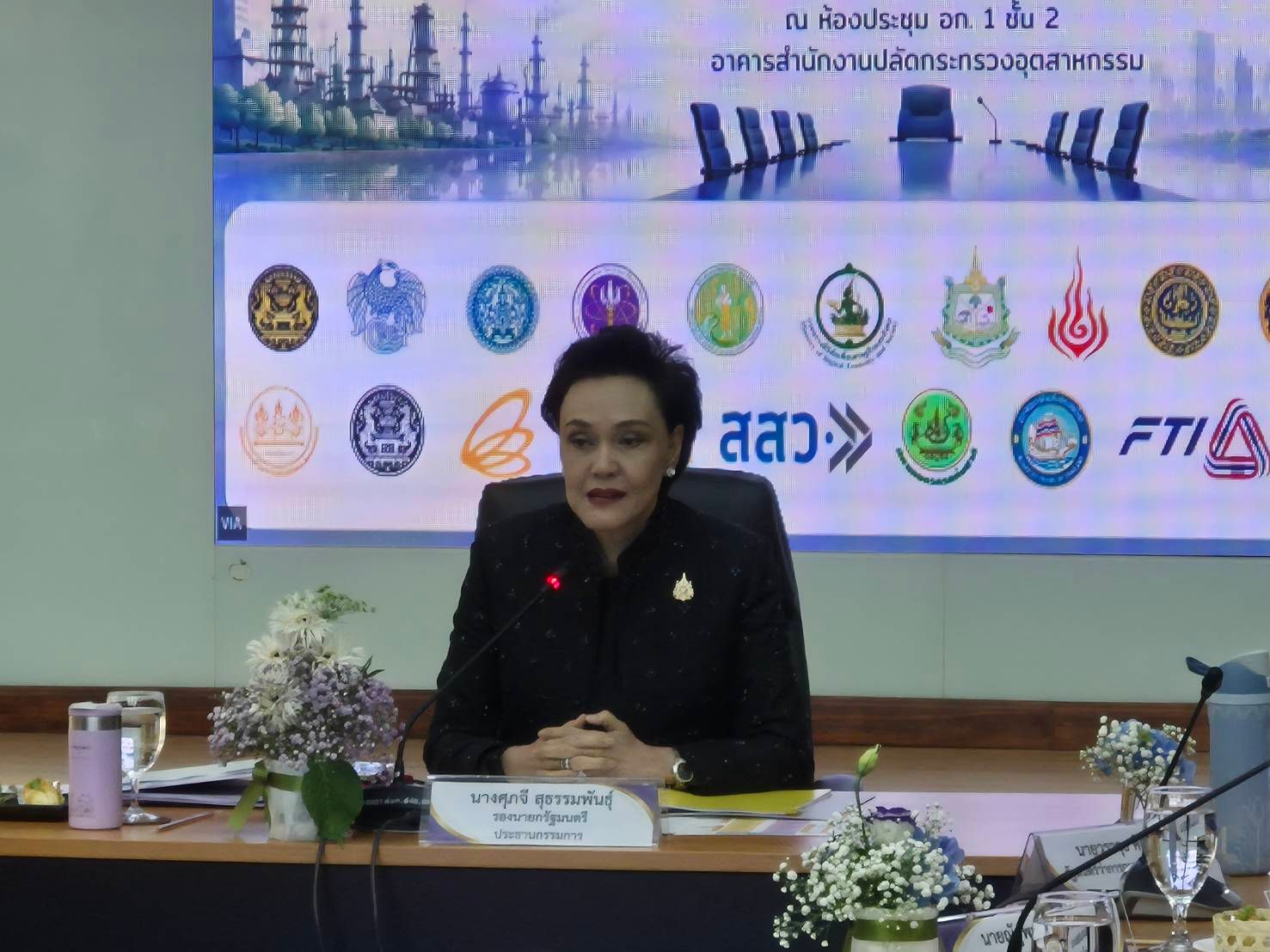 Woman in dark suit at conference table with organizational logos on screen.