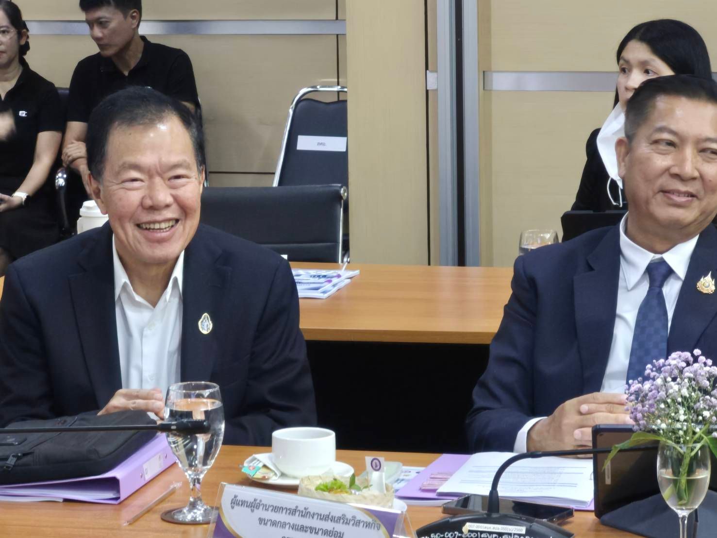 Two men in suits smiling at a conference table with documents and drinks.