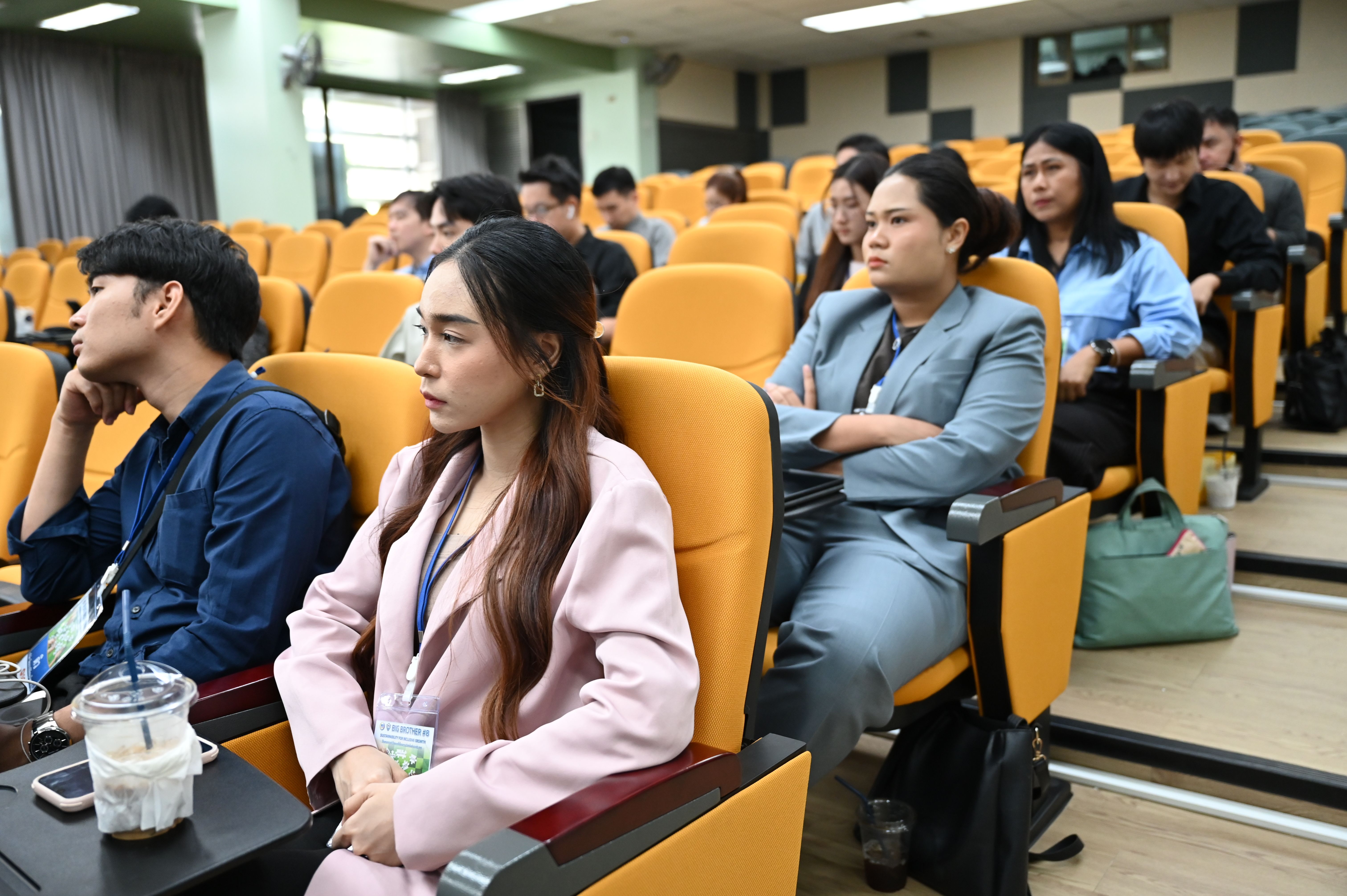 Attendees in a lecture hall intently listen, seated in rows of orange chairs.
