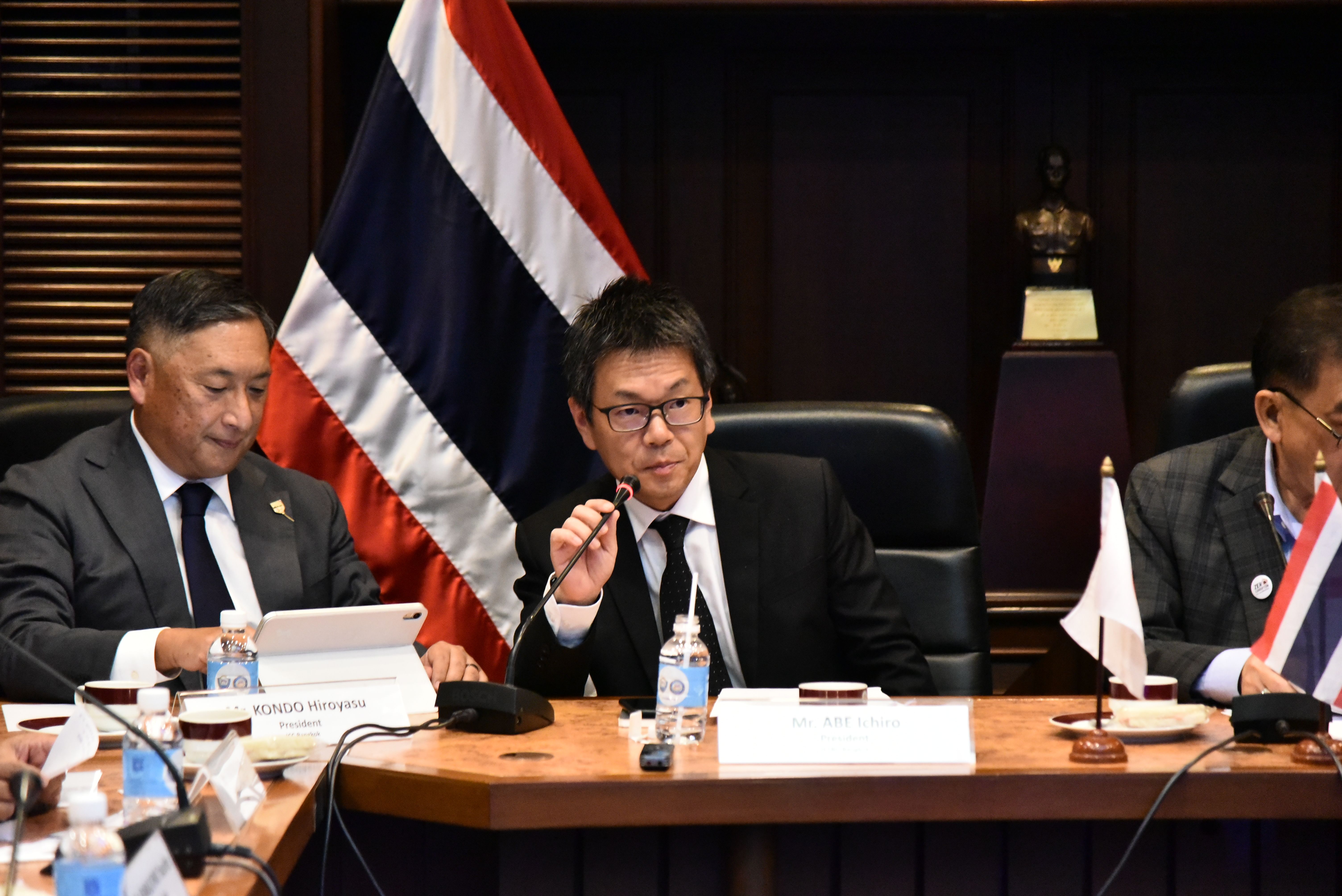 Man speaks into microphone at conference table with Thai and Japanese flags.
