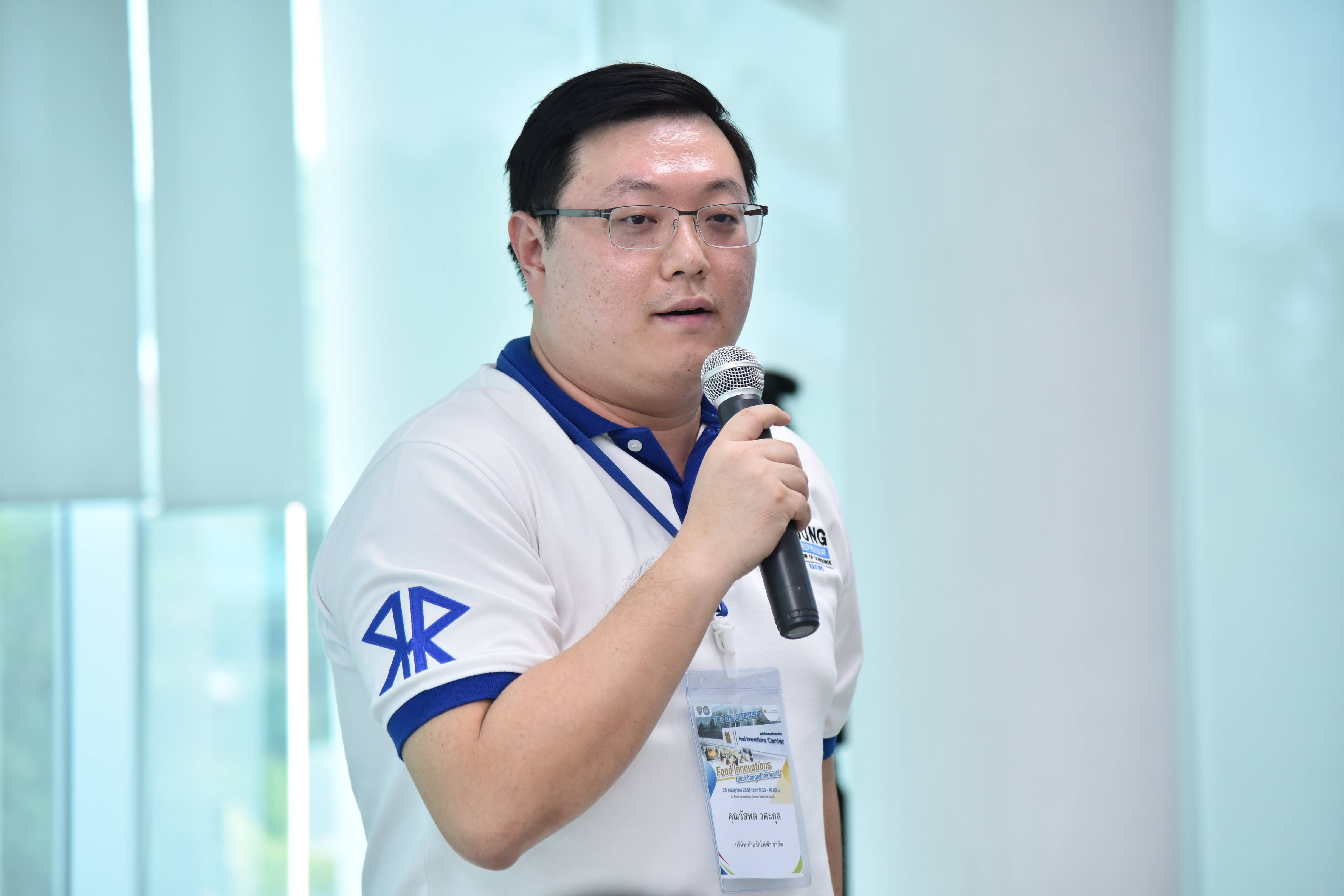 Man in glasses and polo shirt speaking into a microphone at an indoor event.