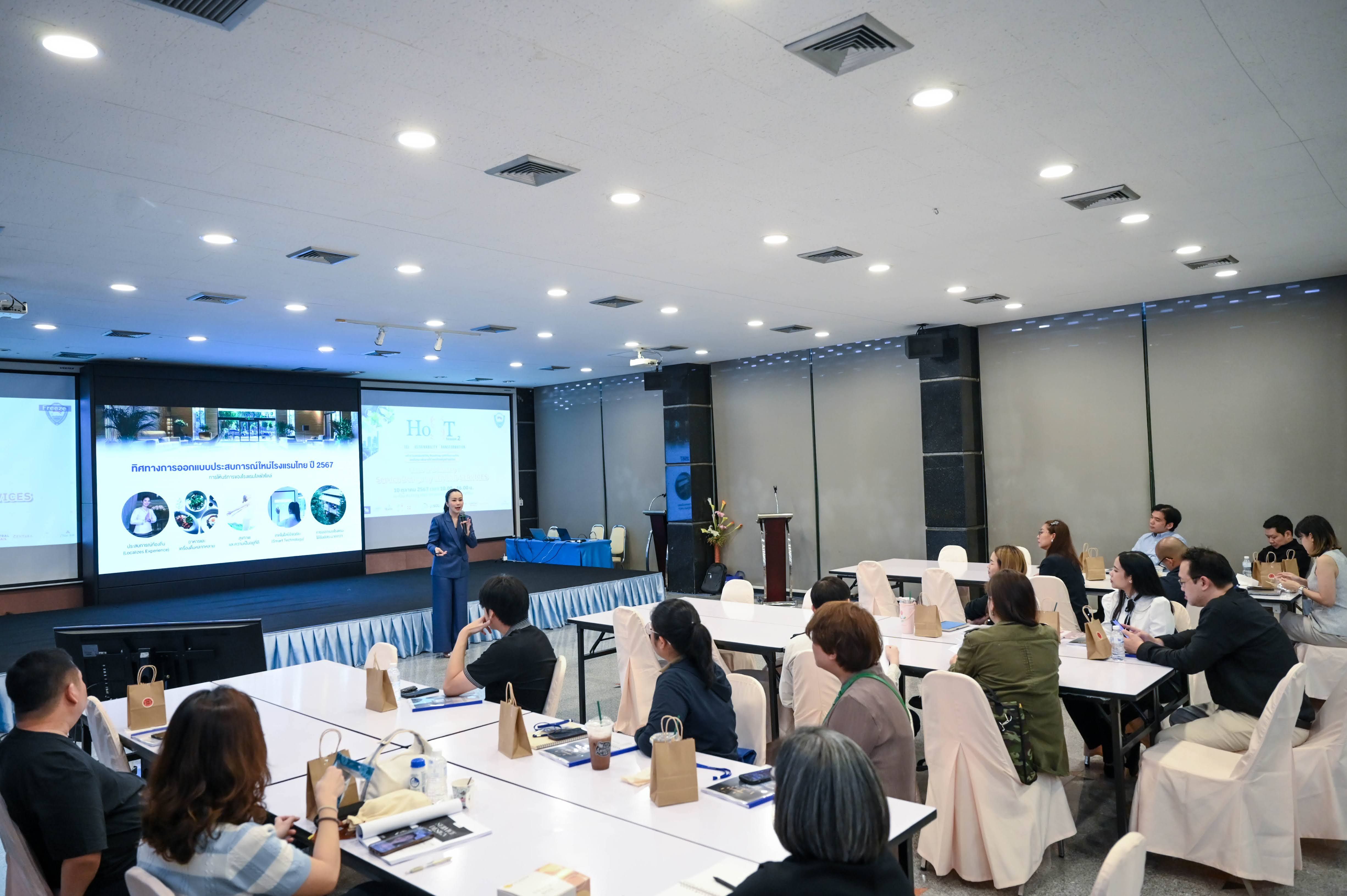 Presenter speaks to an audience in a conference room with a large screen and presentation.