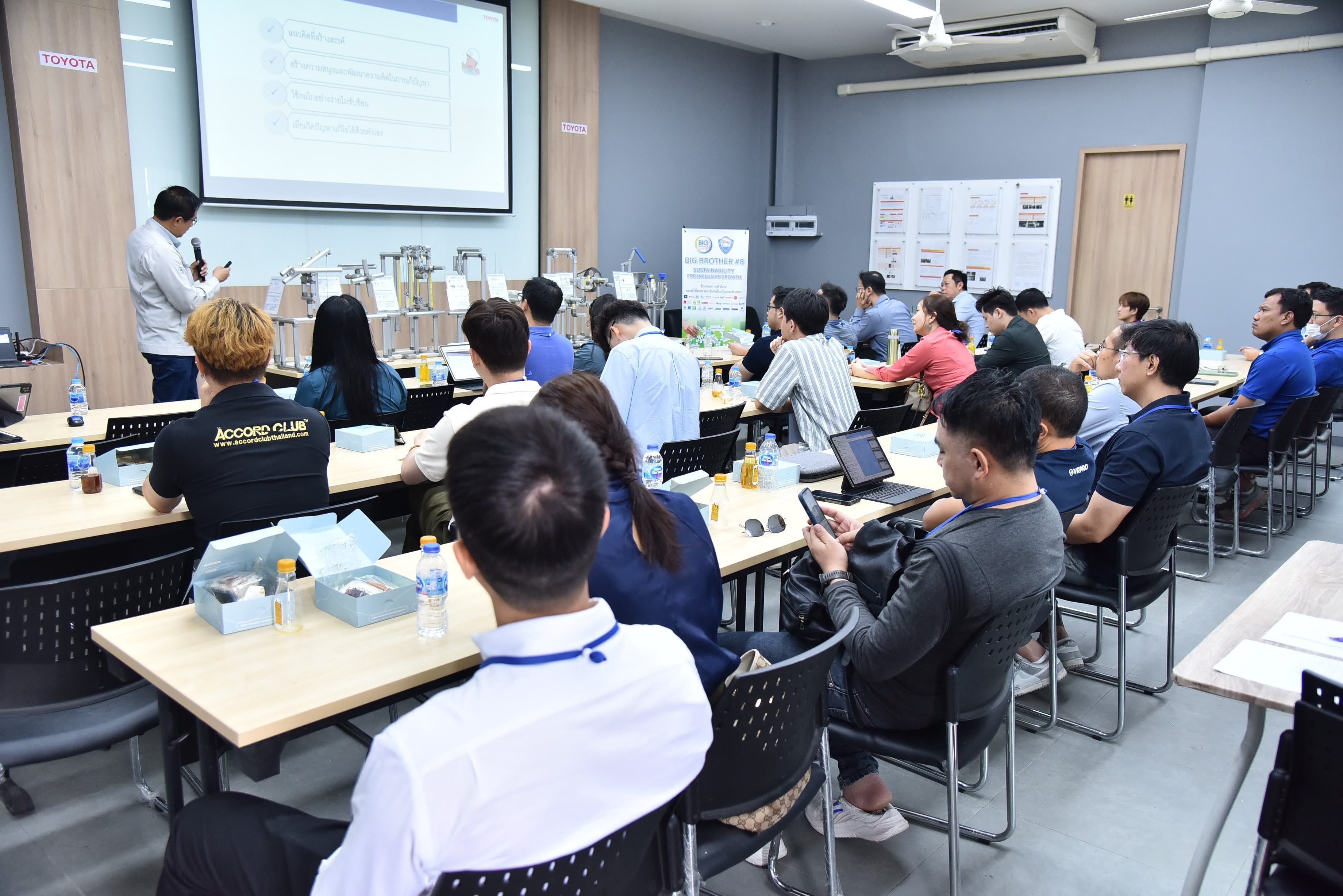 A speaker presents to an engaged audience seated in a classroom setting with a projector screen.