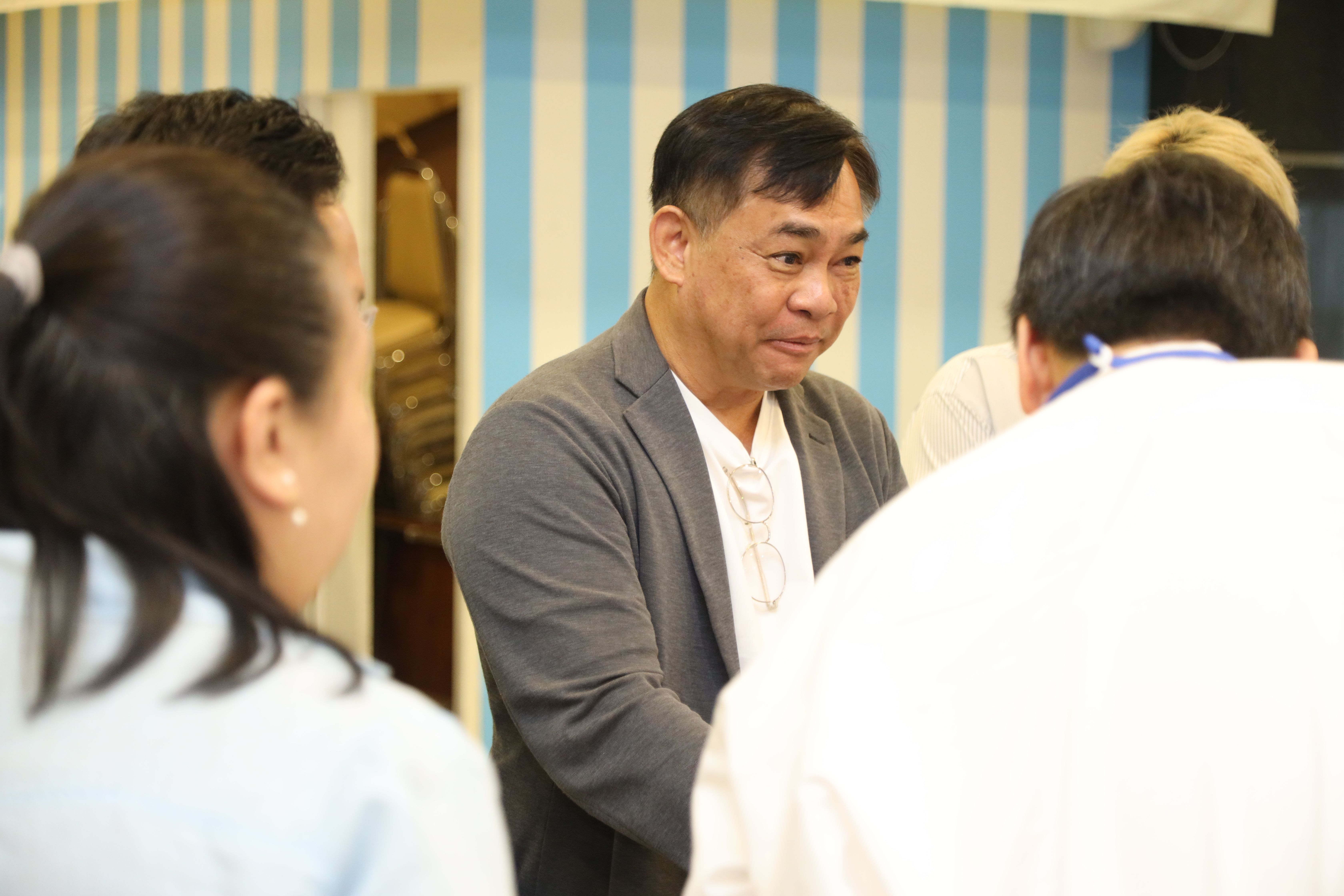 Asian man in grey jacket attentive during an indoor group discussion.