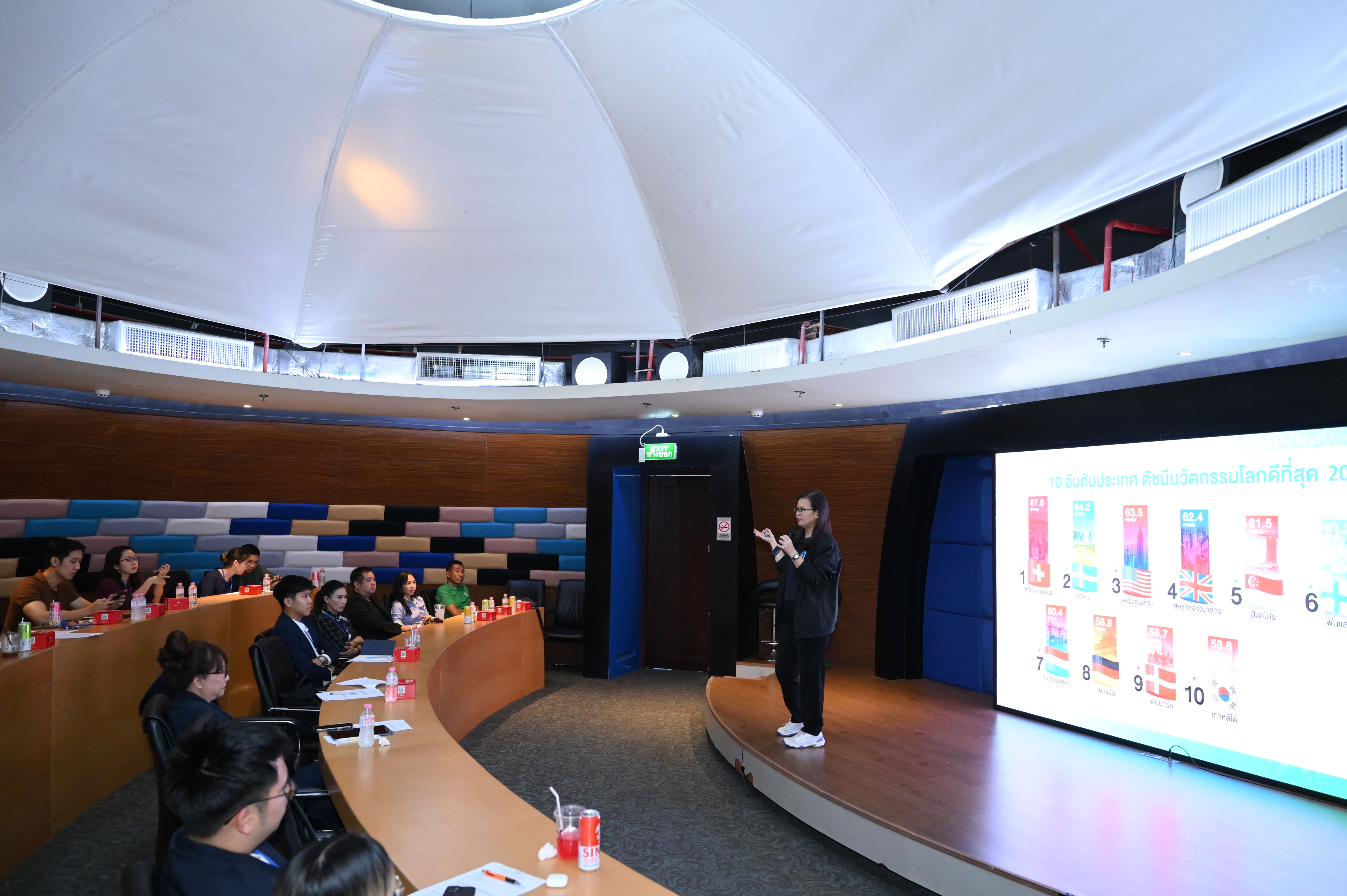 Presenter speaks to an audience in a modern auditorium with a large screen displaying information.