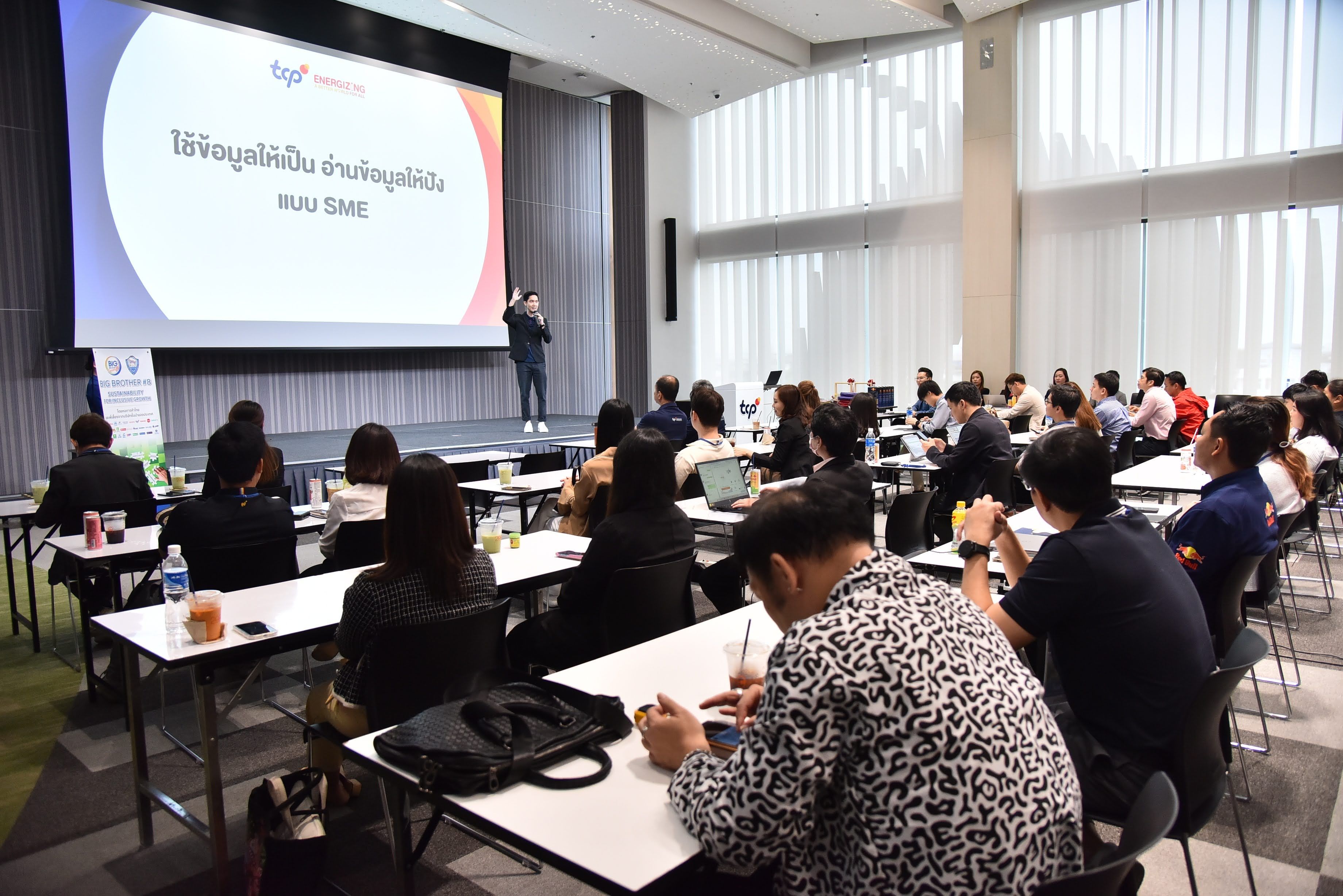 A presenter speaks to an attentive audience in a bright conference room with a large screen.