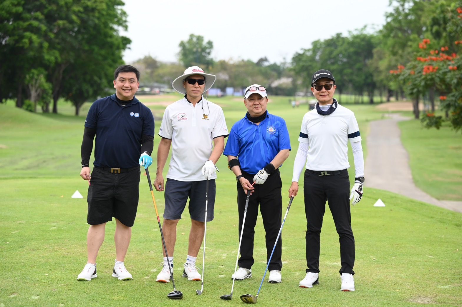 Four men in golf attire pose with clubs on a sunny green golf course.