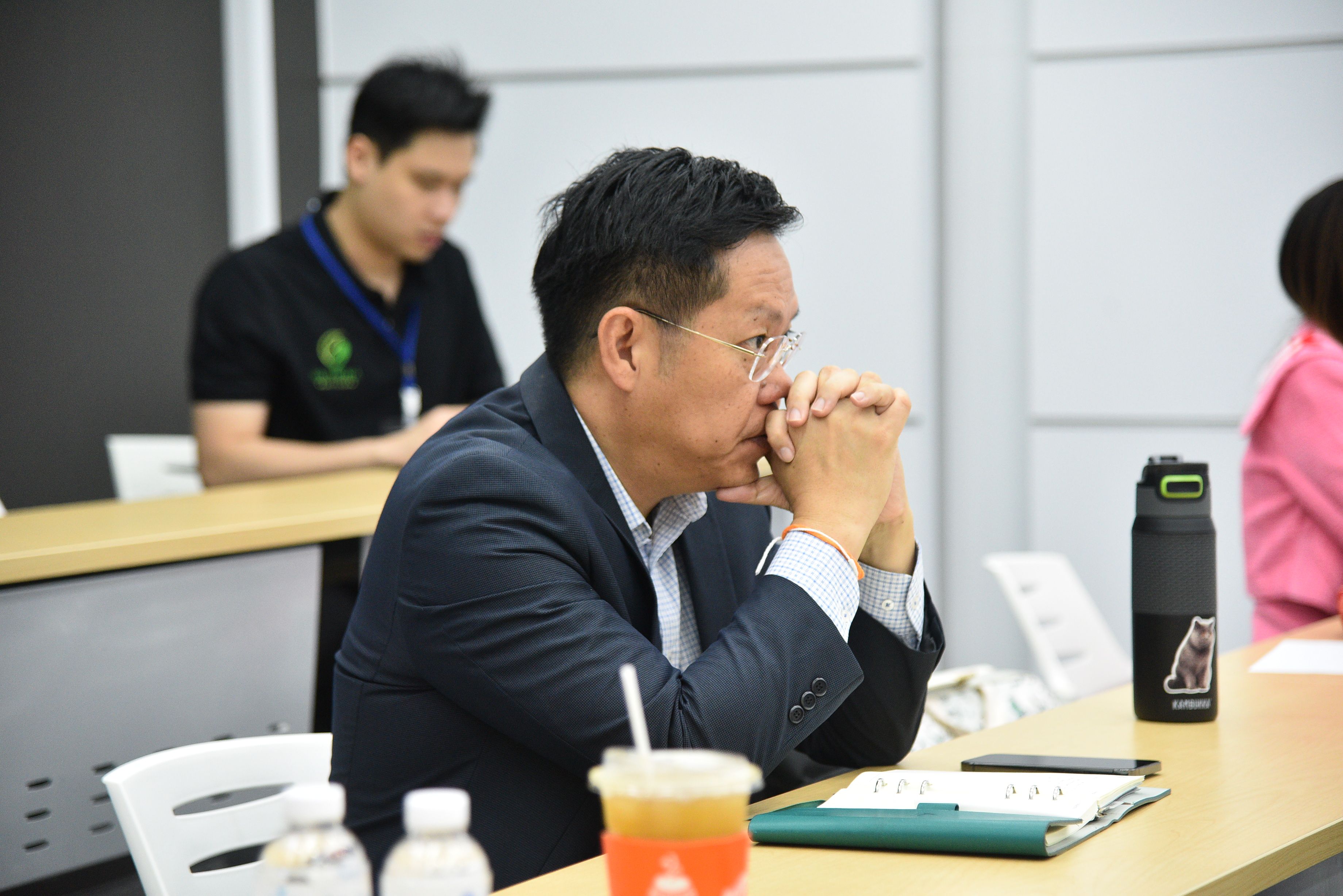 A man in a suit attentively listens with hands clasped in a conference setting.