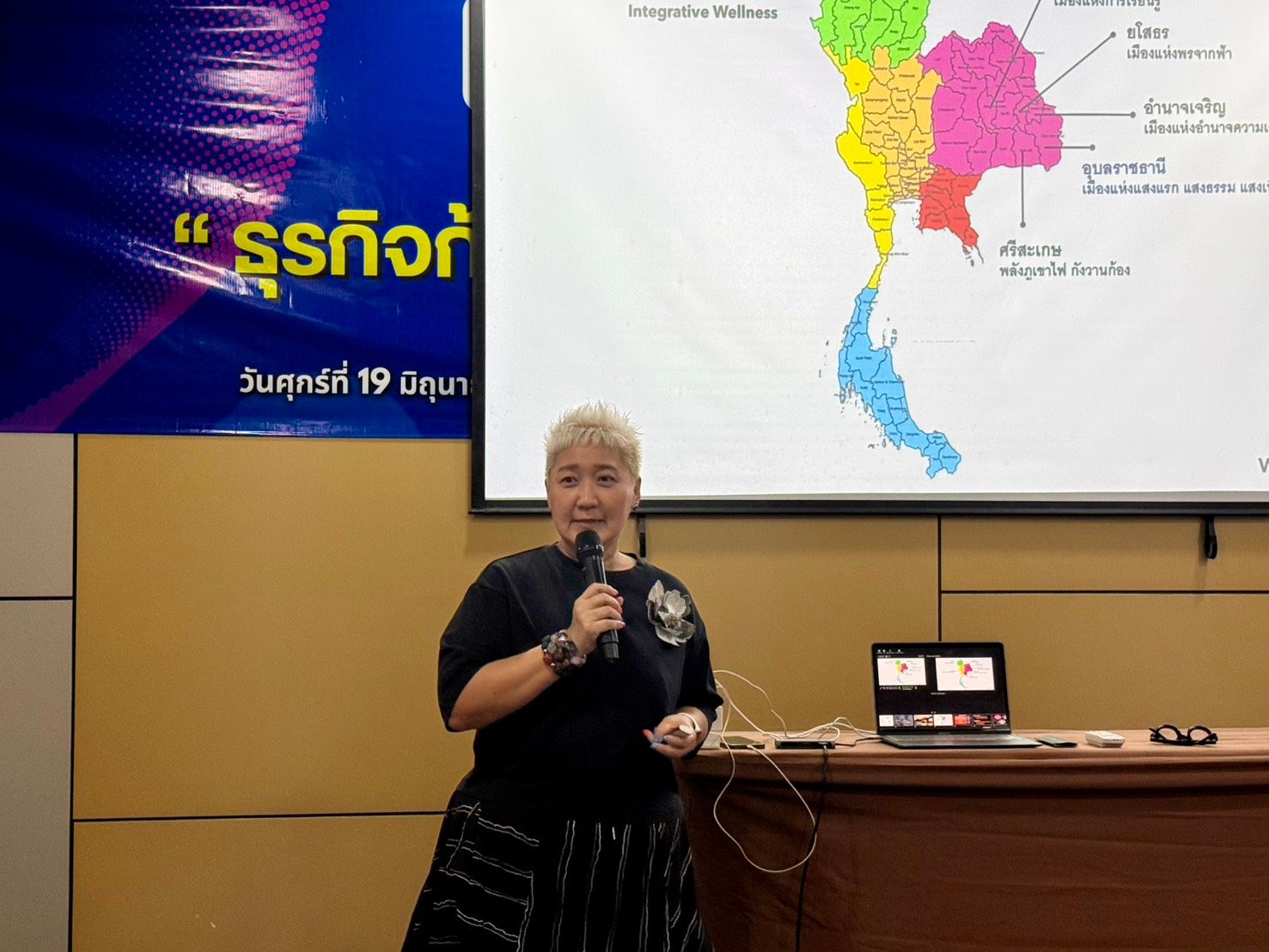 Speaker with white hair and black top presenting in front of a map.