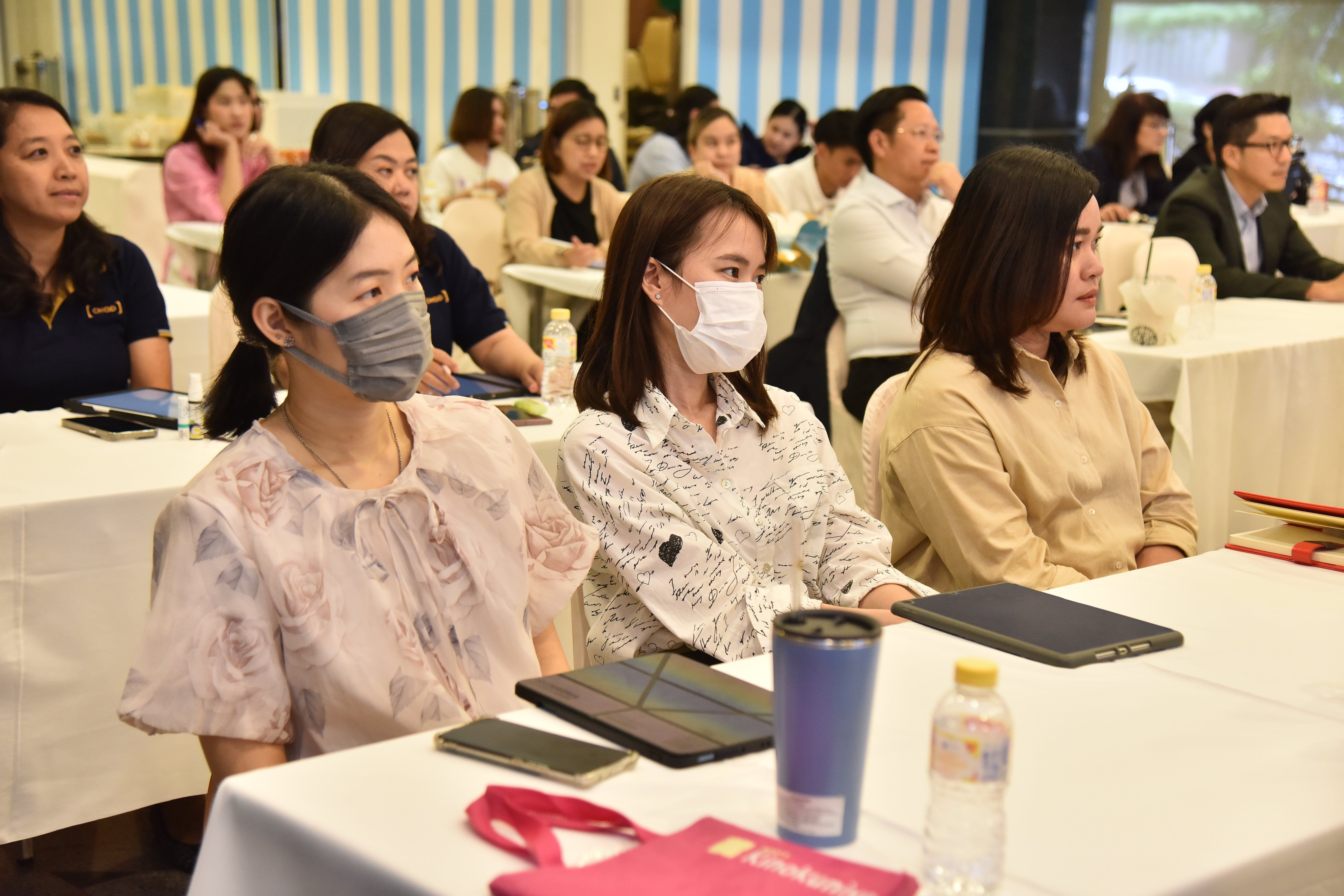 Attendees, some wearing masks, listen attentively at a conference in a brightly lit room.