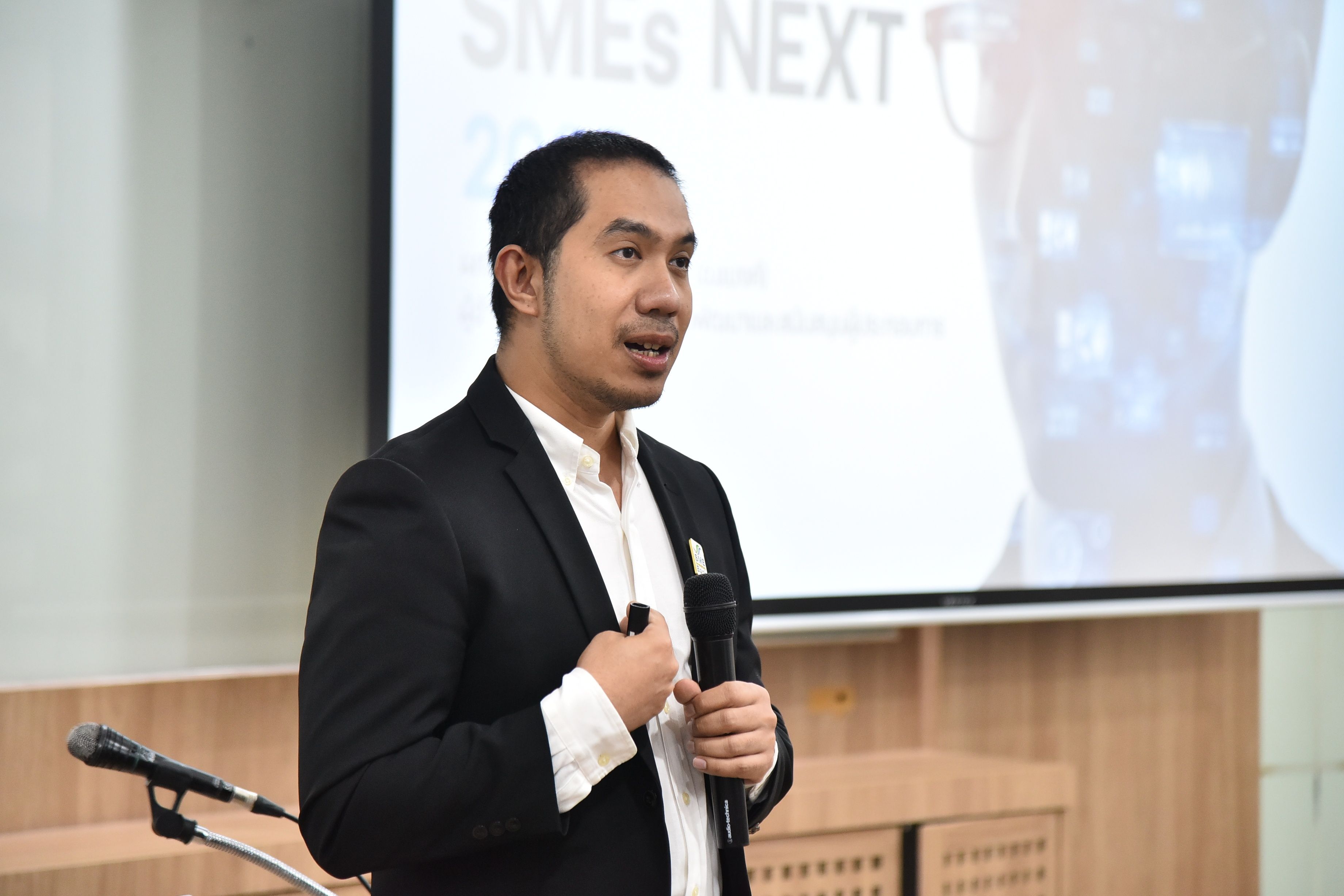 Man in black suit speaks into microphone at a conference with 'SMEs NEXT' on screen.