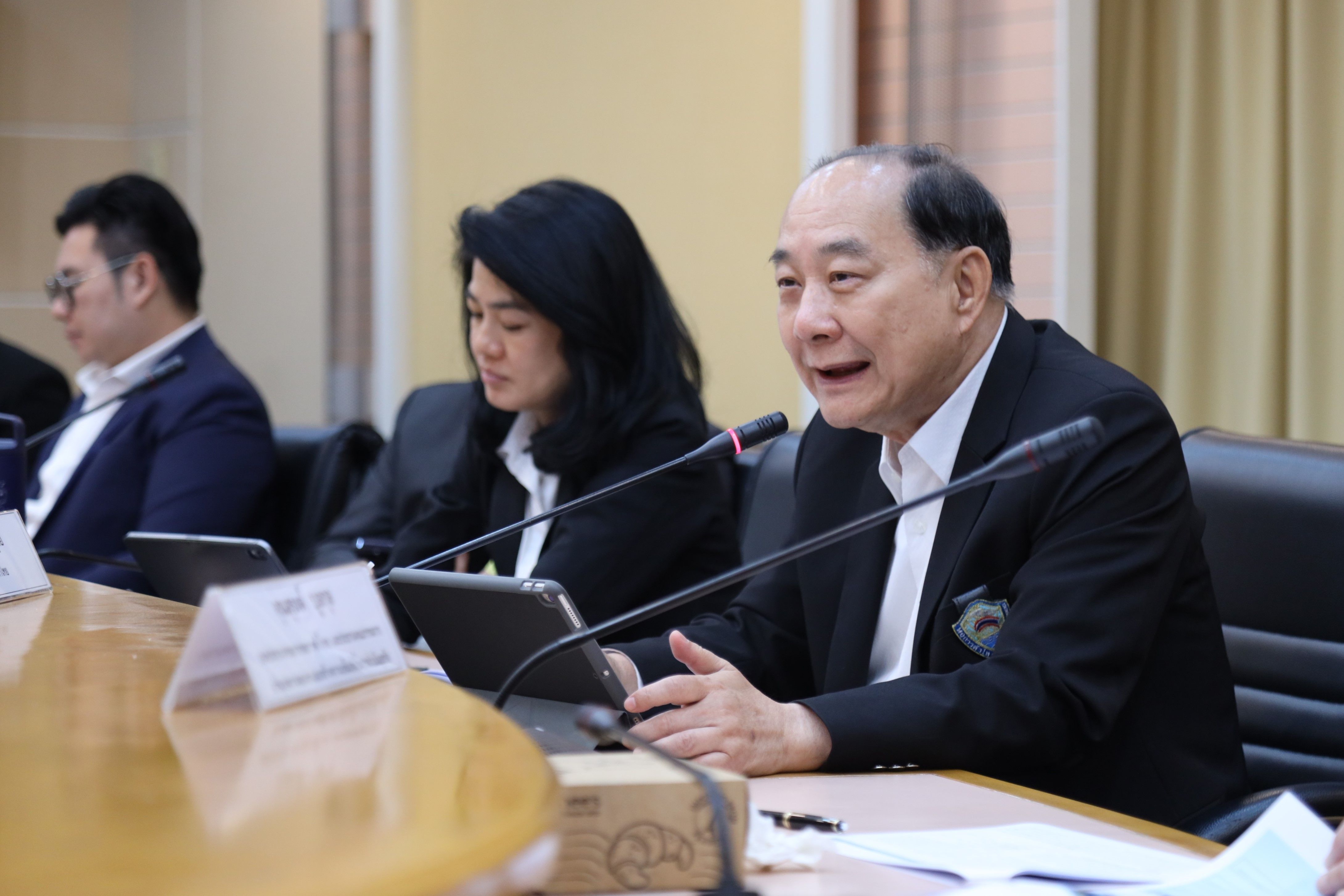 A man in a dark suit speaks at a meeting table with colleagues.