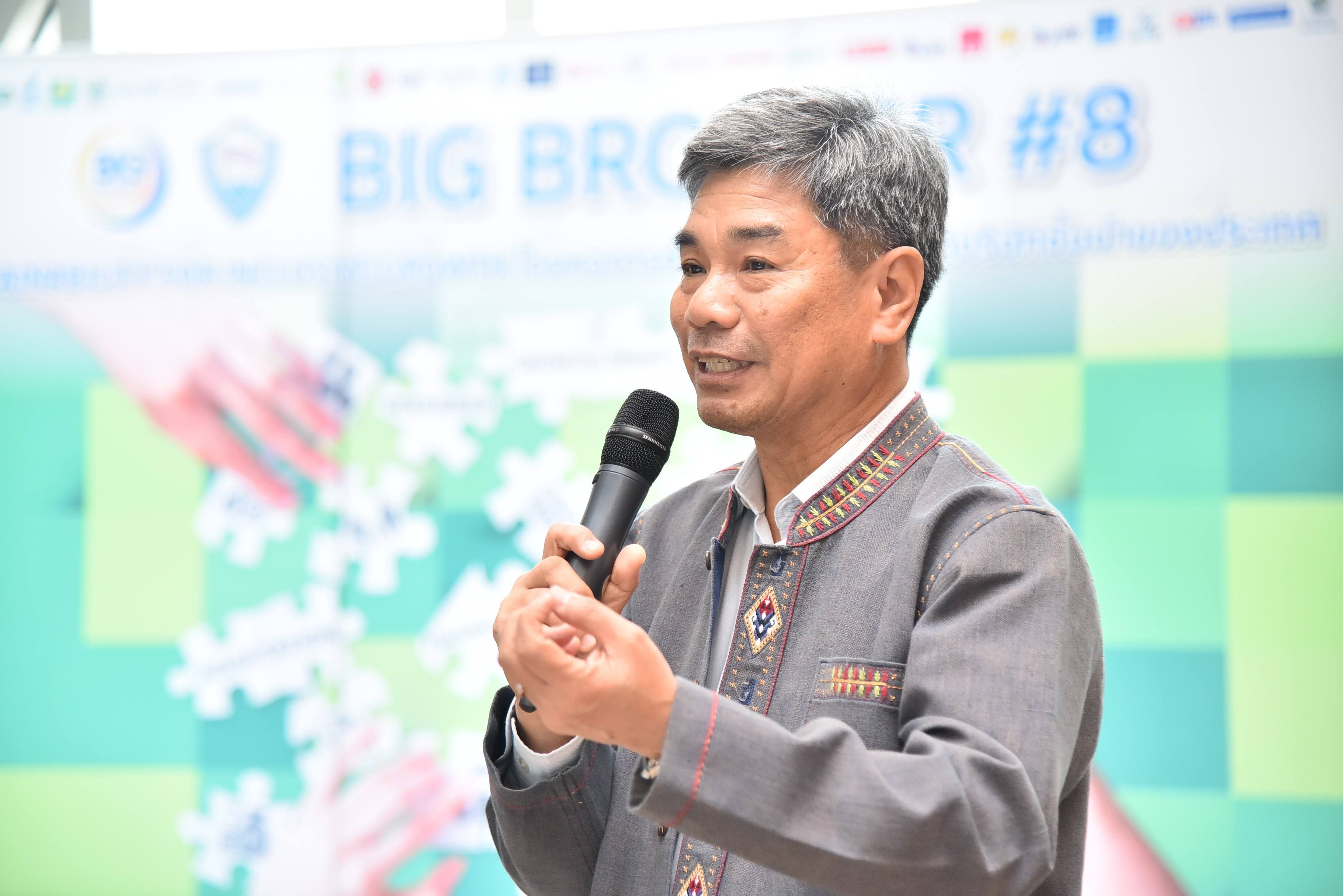 A man in a patterned shirt speaks into a microphone at an indoor event.