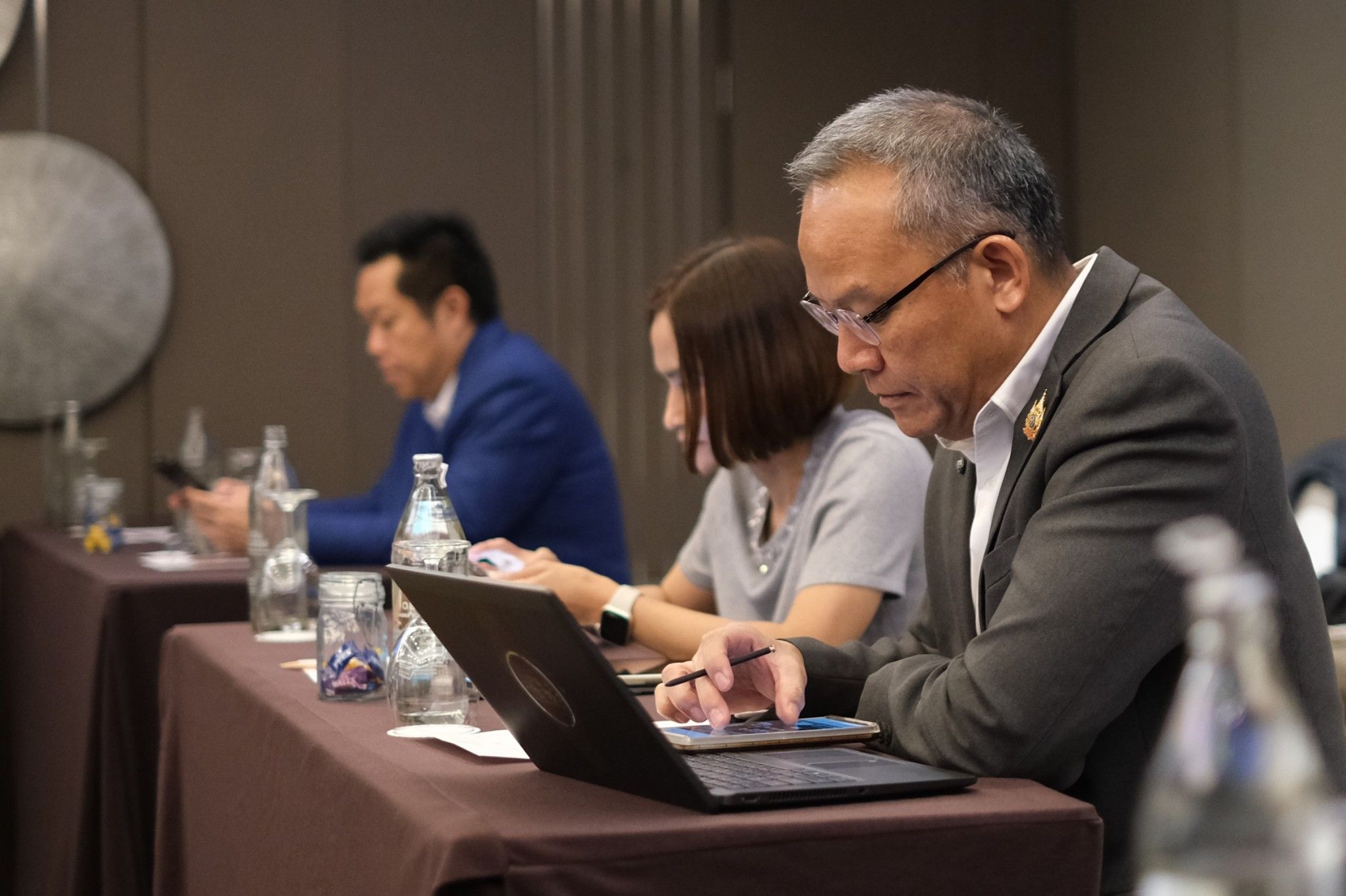 A man in glasses uses a laptop at a conference table with two other attendees.