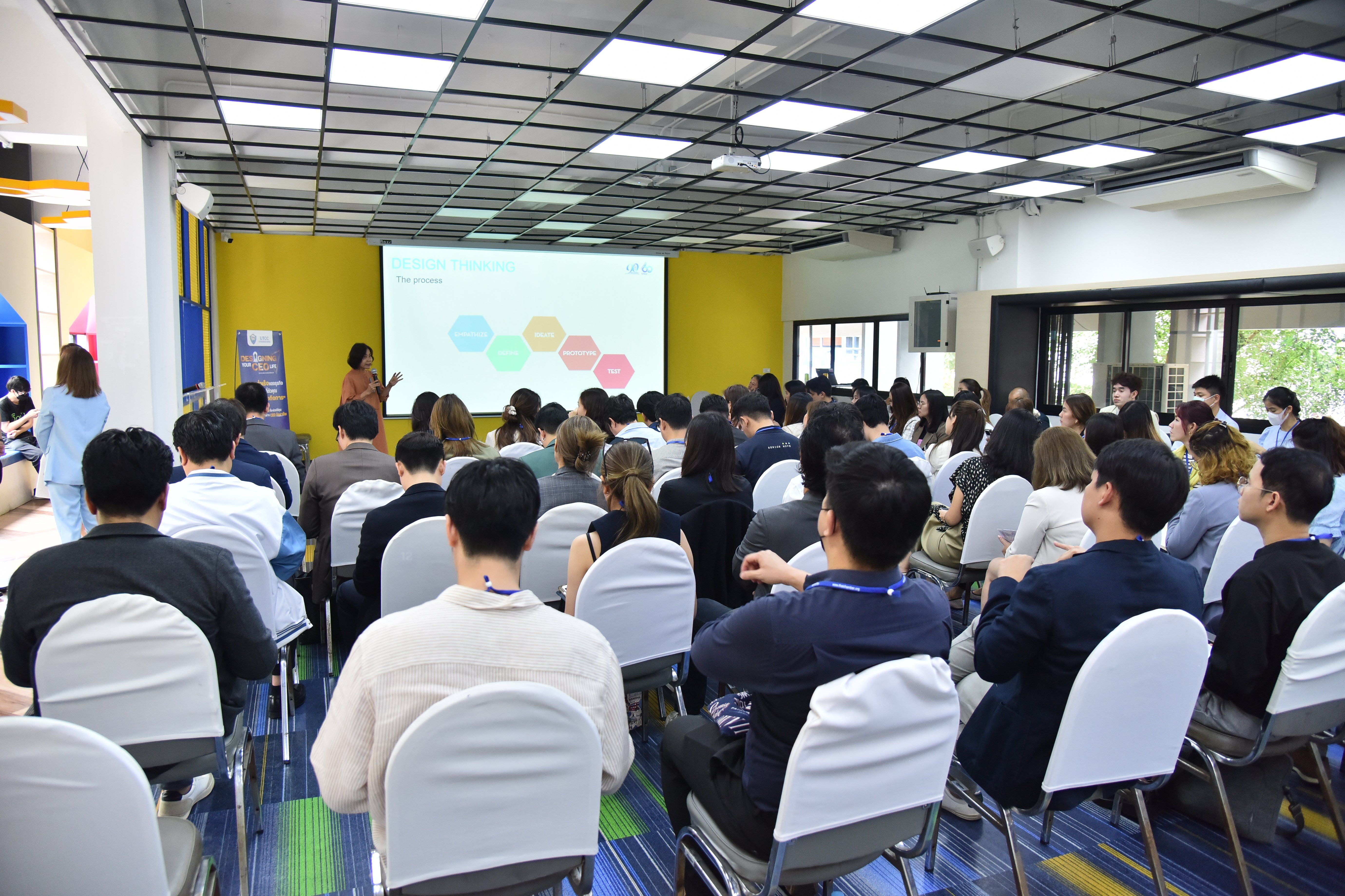 Audience watches a presenter with a colorful diagram on screen in a modern seminar room.