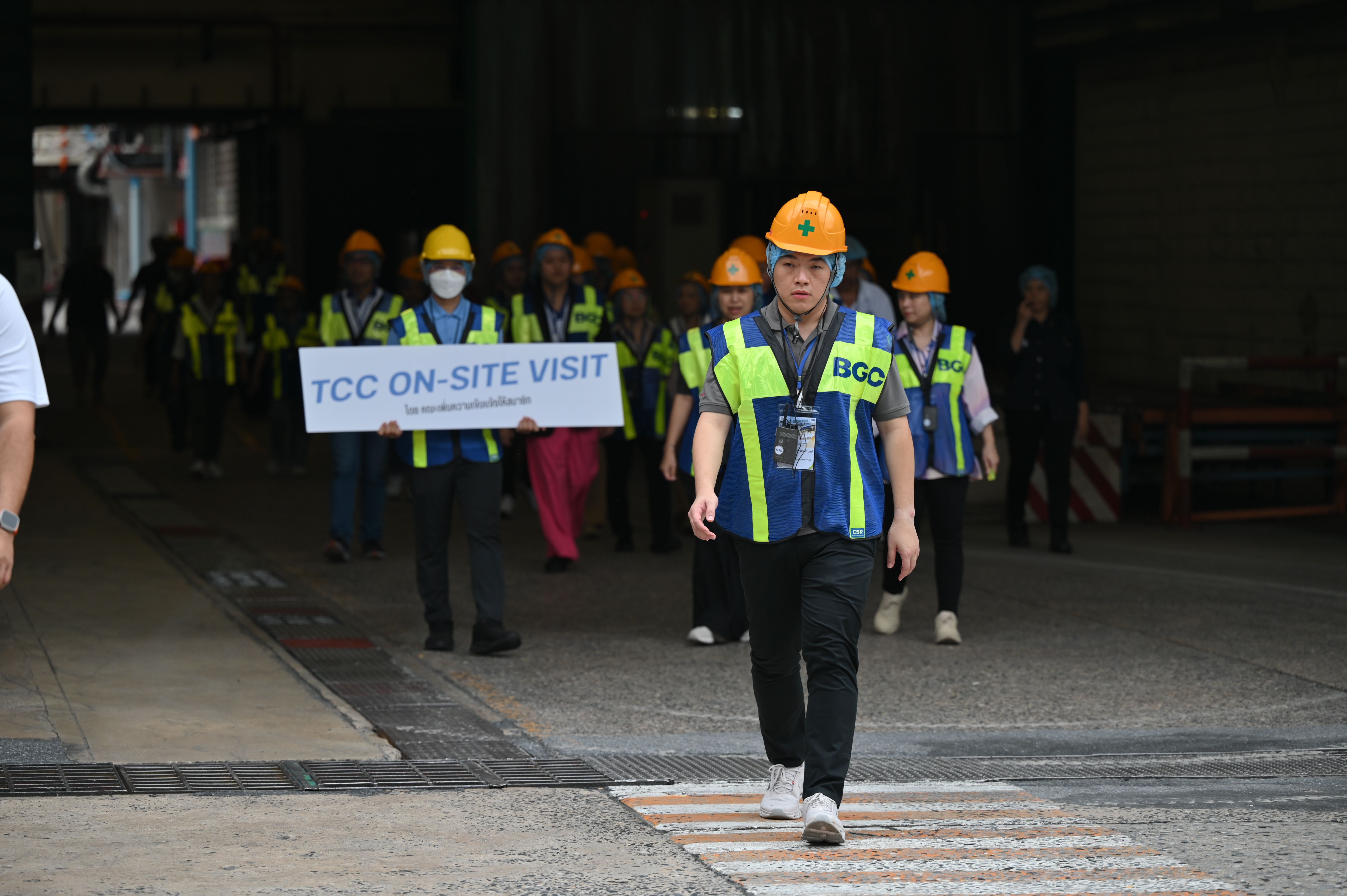 Group wearing hard hats and safety vests on site visit, one holds 'TCC ON-SITE VISIT' sign.