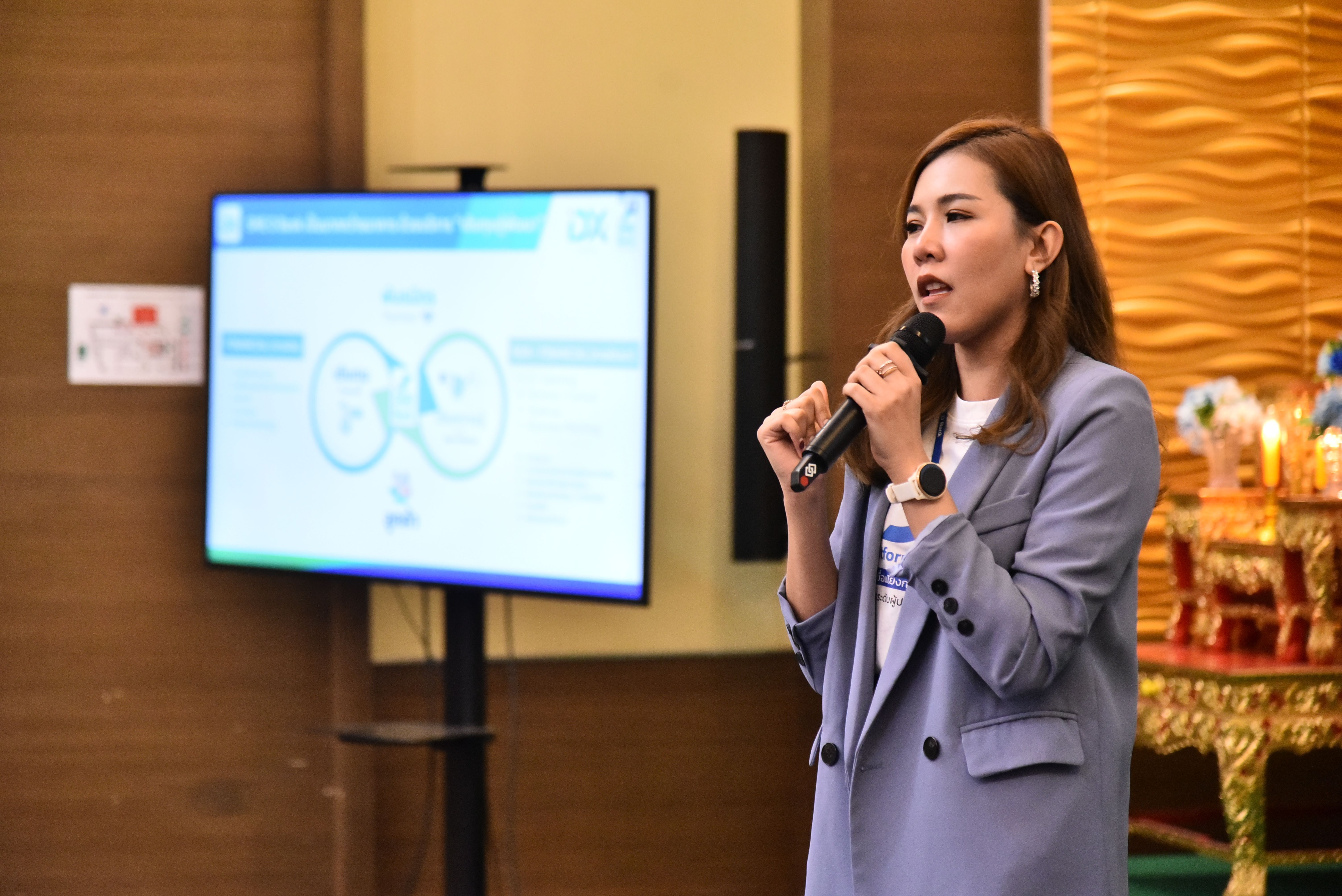 Woman in a blazer speaking into a microphone during a presentation with a diagram on screen.
