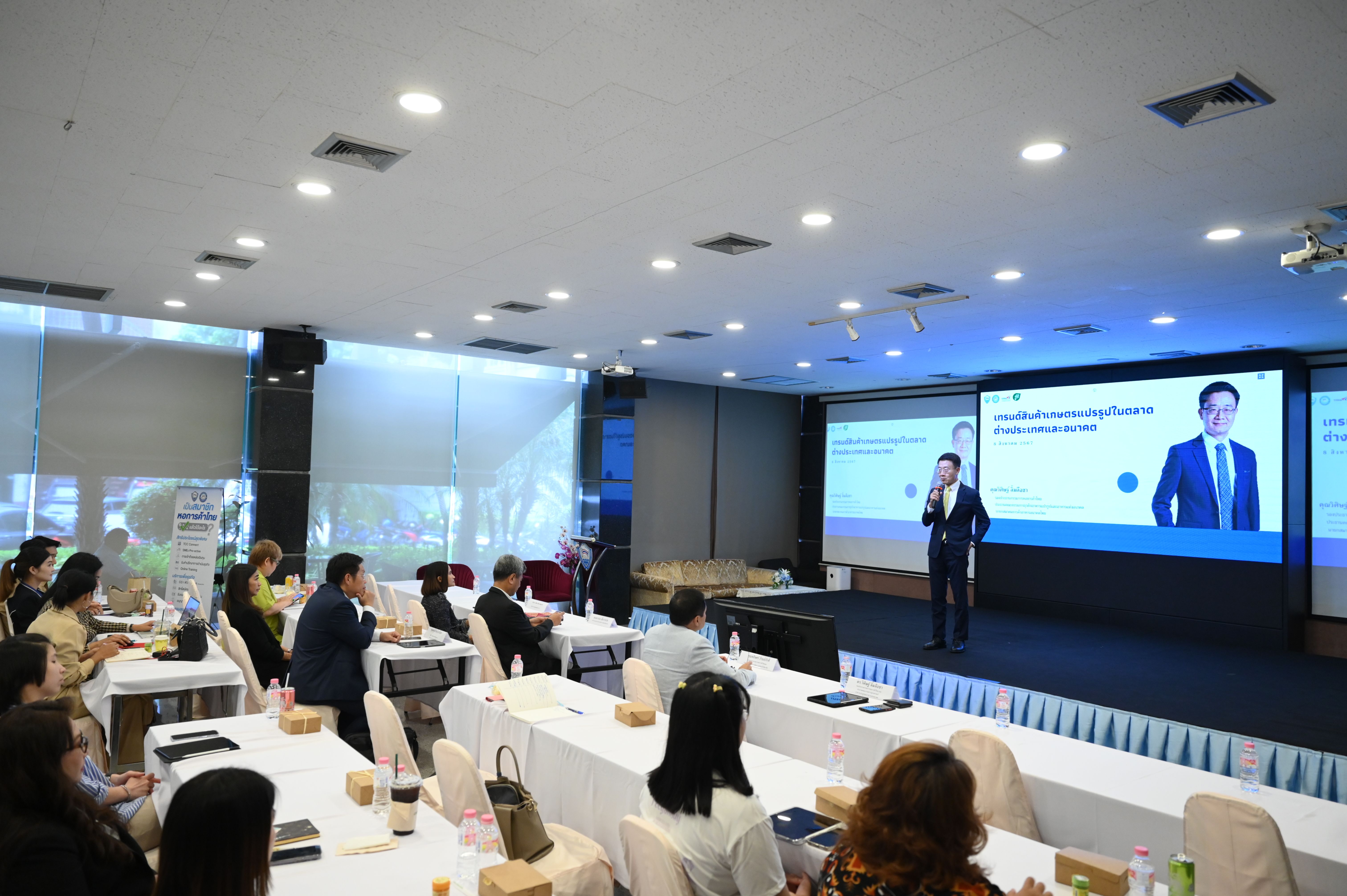 A man presents on stage to a seated audience in a bright conference room.