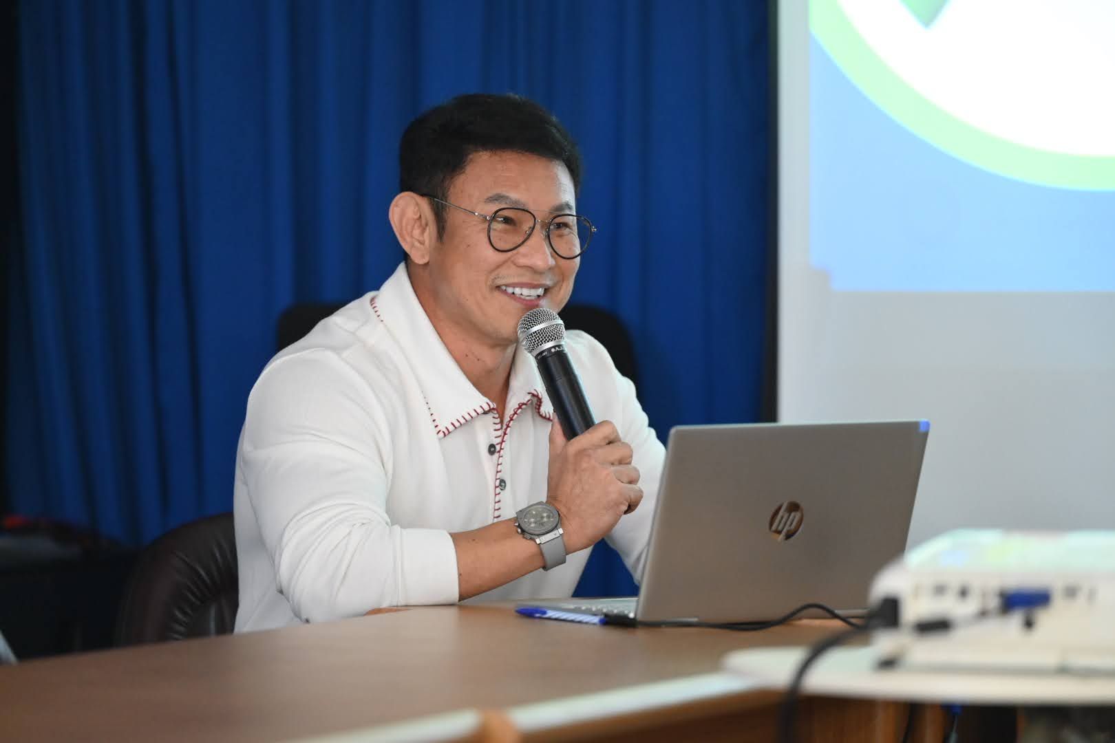 Man in glasses speaking into microphone during presentation, smiling beside laptop and projector.