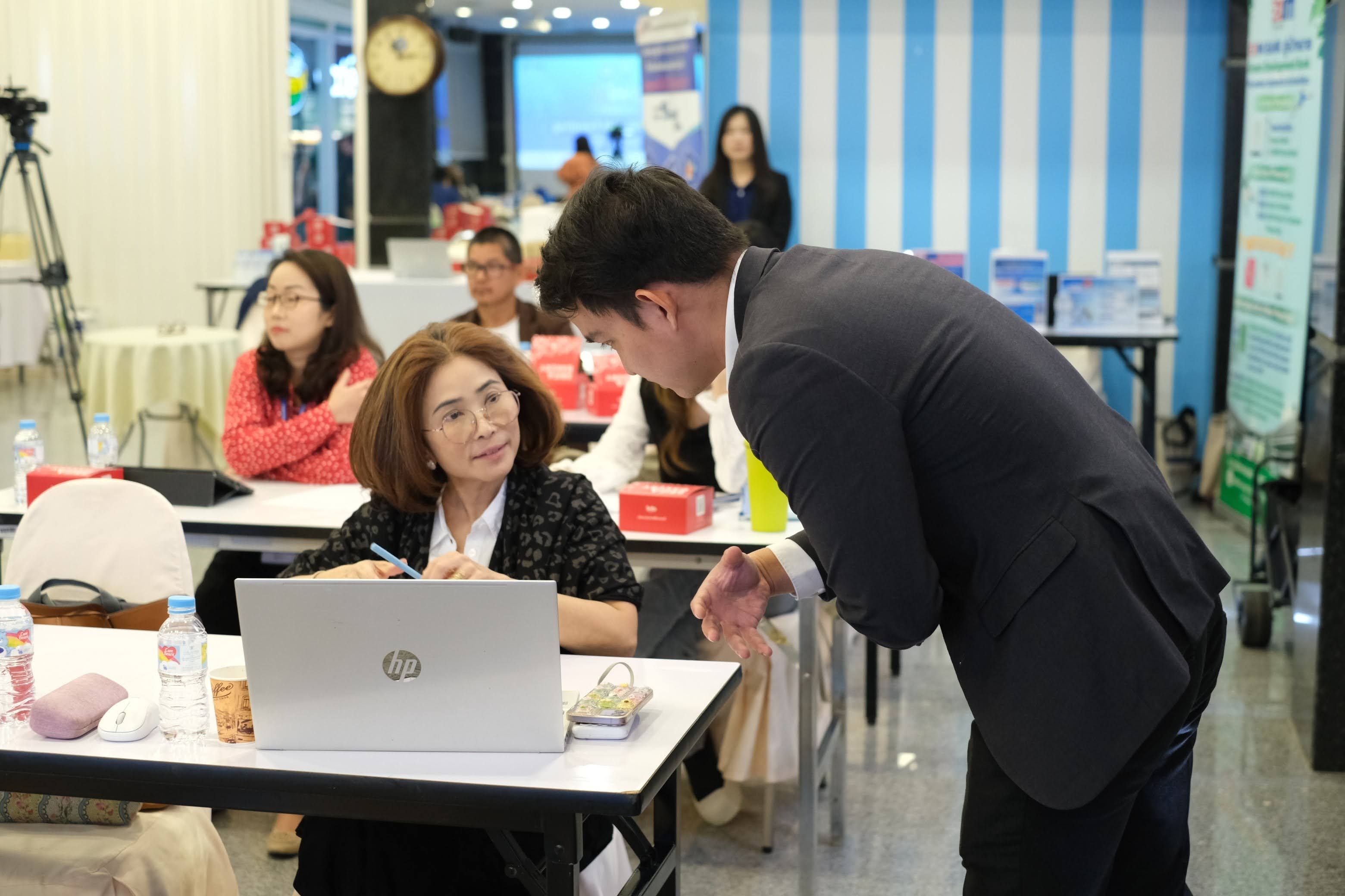 A man talks to a woman at a desk with a laptop in a classroom.