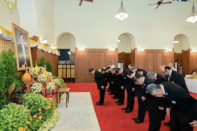 Men in black suits bow before a decorated altar displaying a framed portrait.