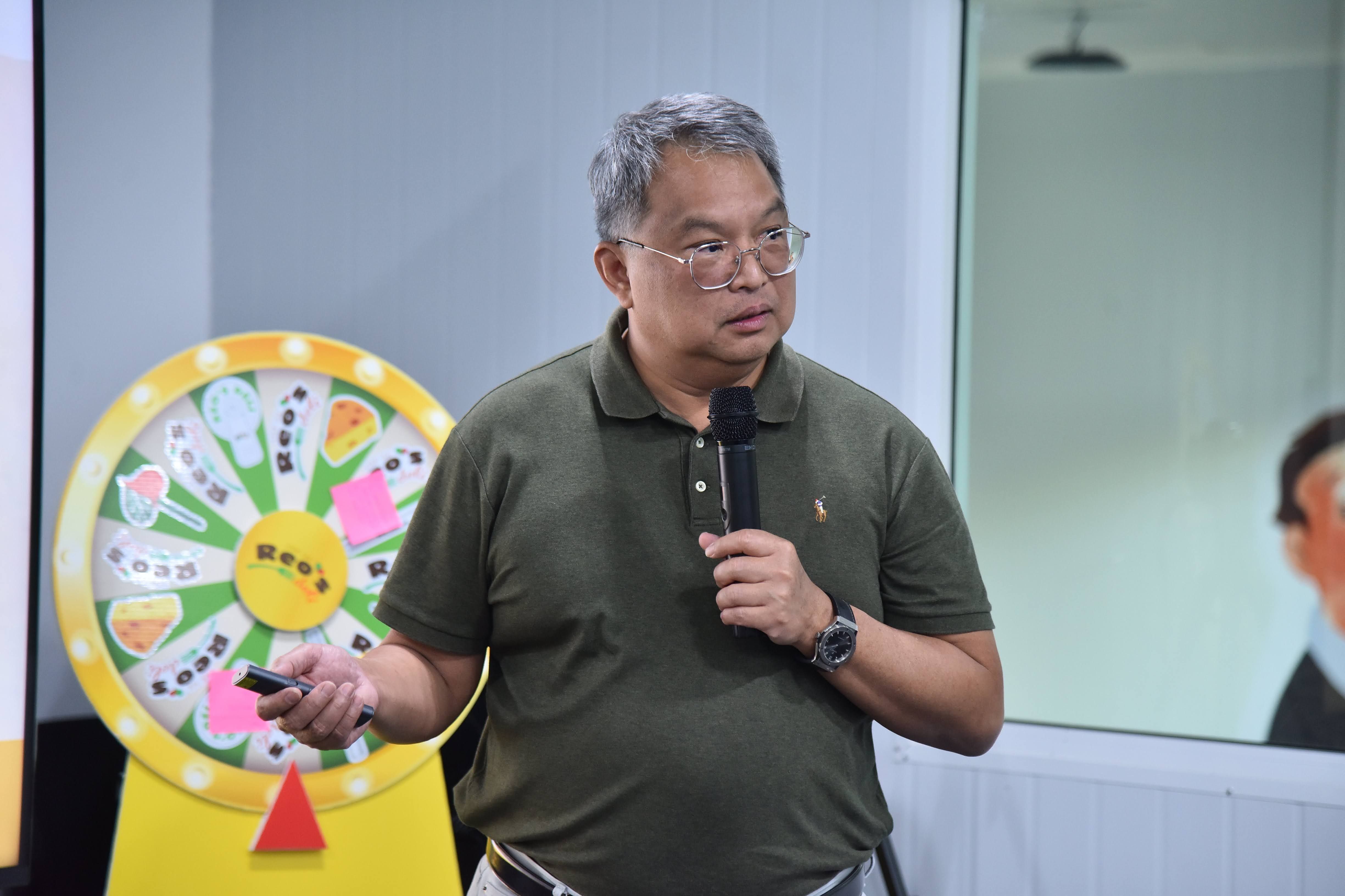 Man in glasses, polo shirt, speaks into microphone, holds clicker, spinning wheel behind him.