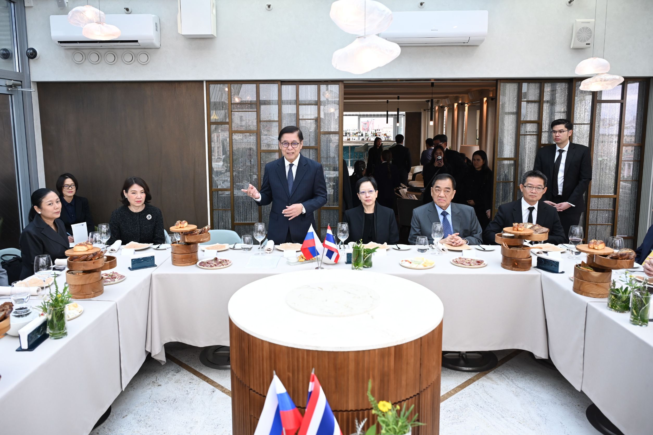 Man in a suit speaking at a formal table with attendees and flags.