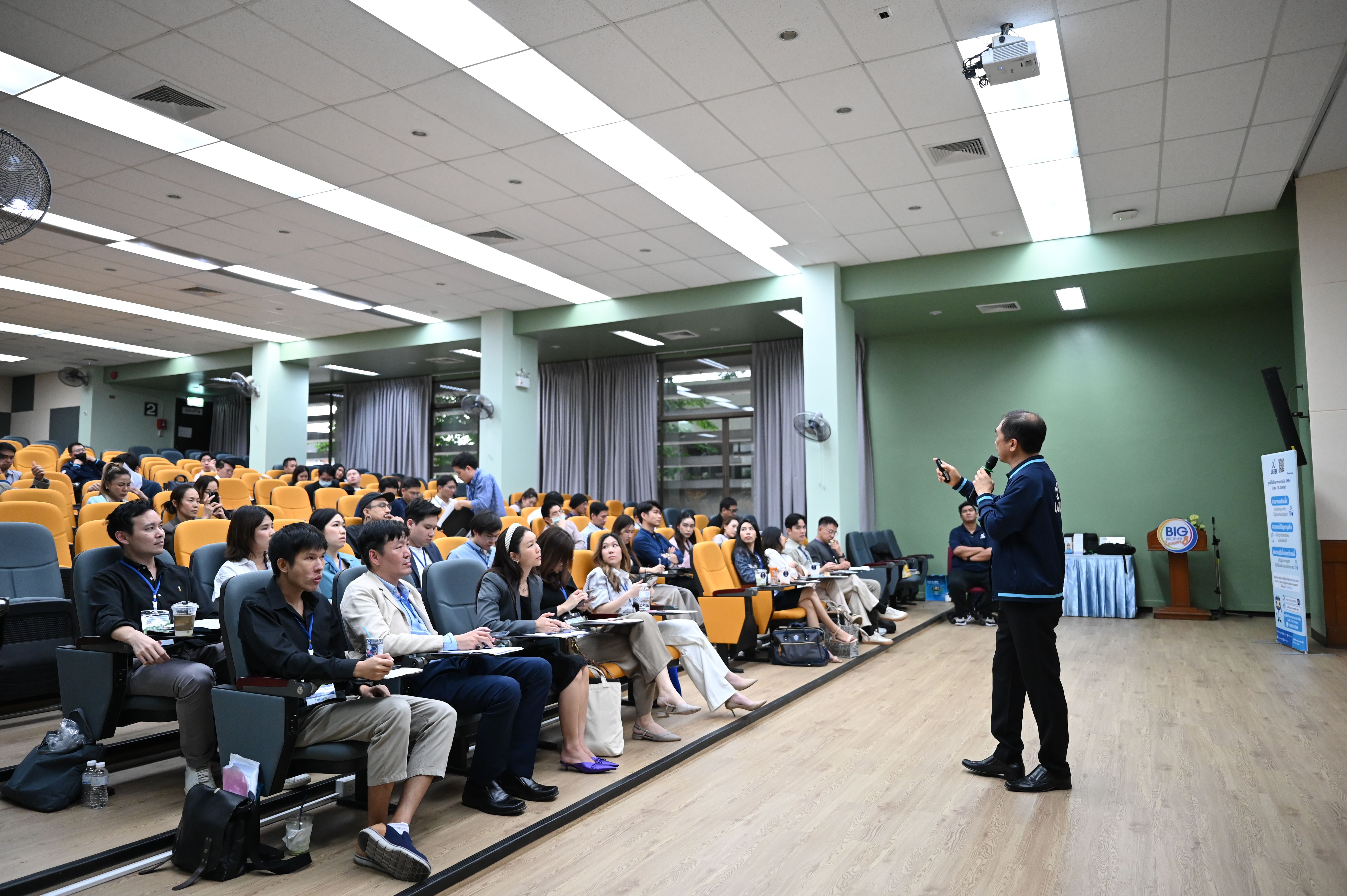 A speaker addresses a large audience seated in rows in a modern lecture hall.