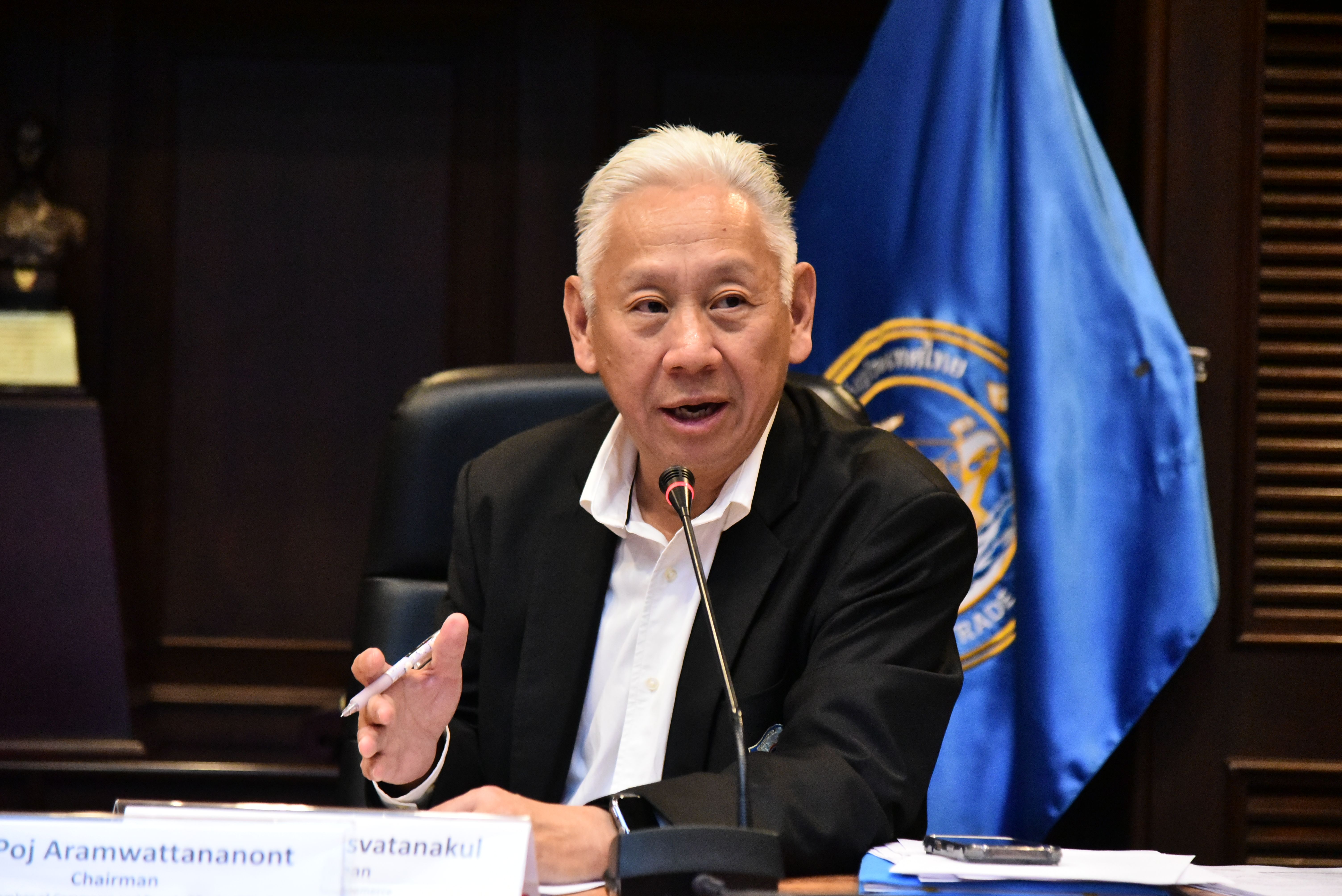 Elderly man with white hair speaking at a table with a blue flag.
