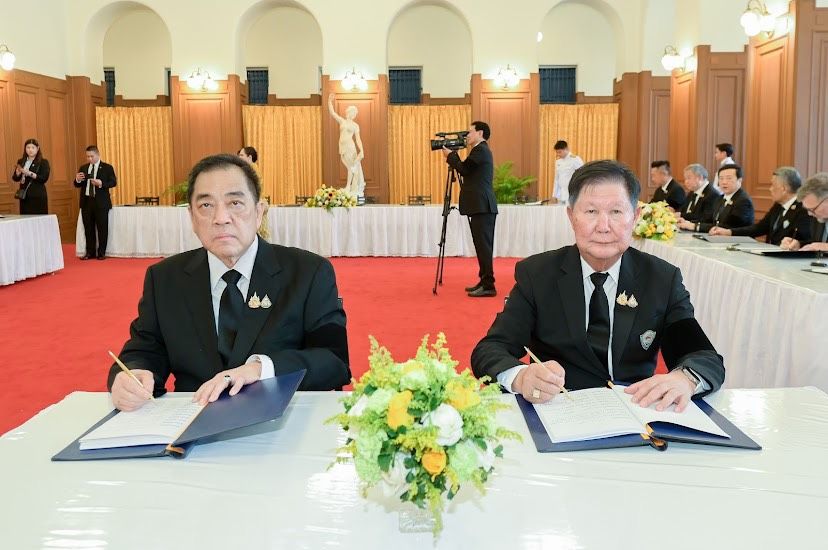 Two men in formal black suits sign documents at a large table in a grand room.